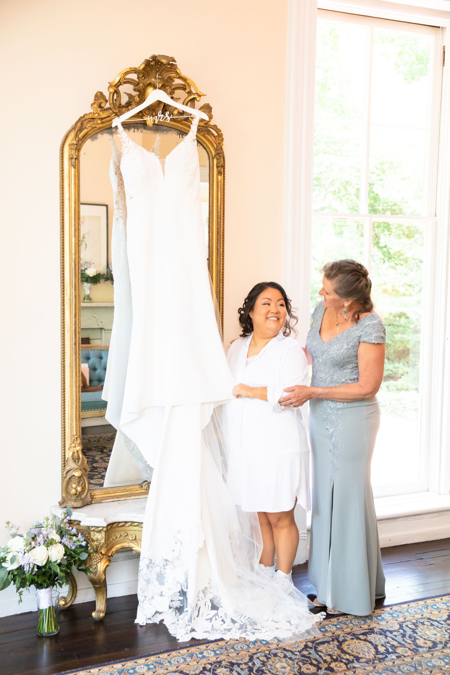 People in a bright room helping with wedding dress preparations near ornate gold mirrors.