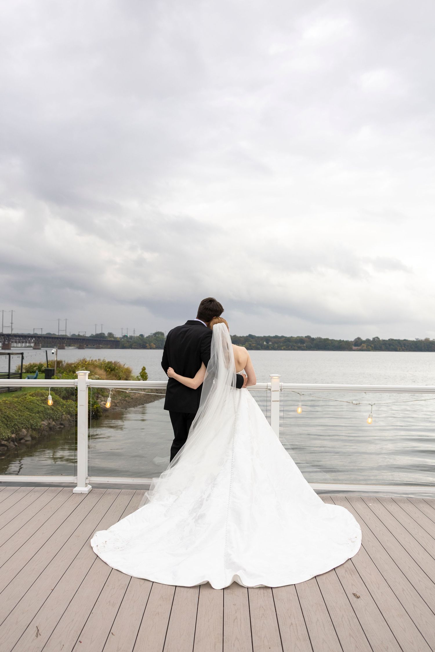 Bride and groom stand embracing on a waterfront deck, her long white gown and veil trailing behind under cloudy skies.