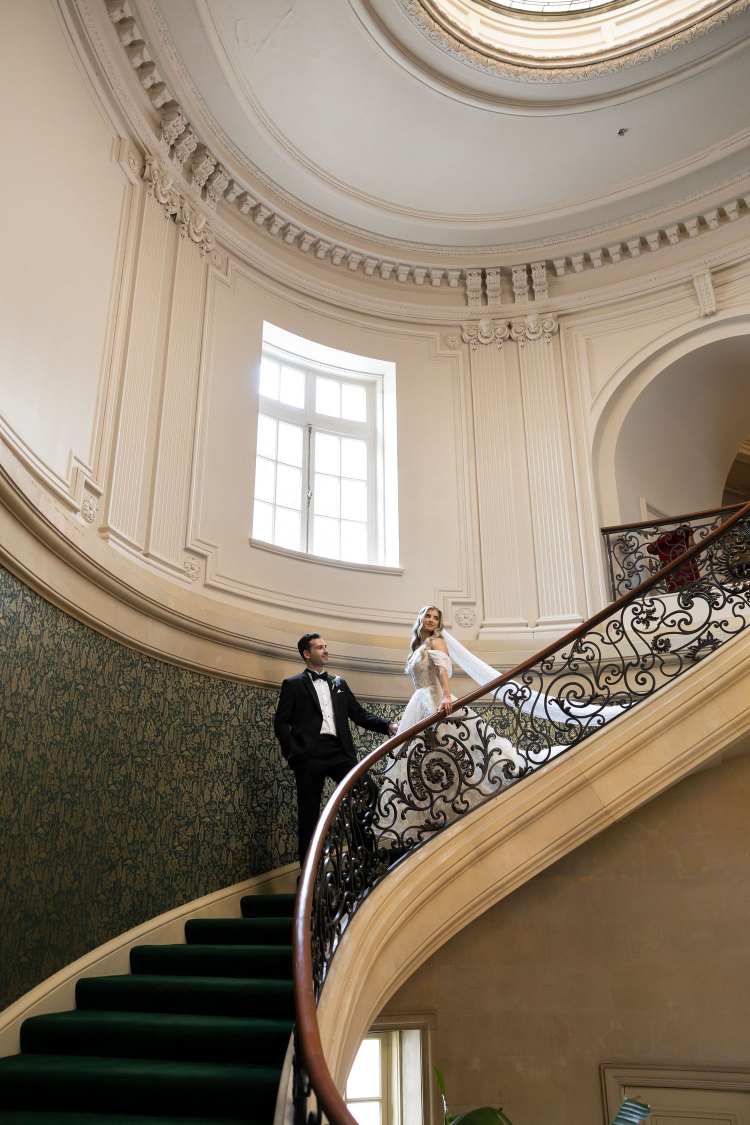 Bride and groom stand on elegant curved staircase with ornate iron railing in a grand classical ballroom venue.