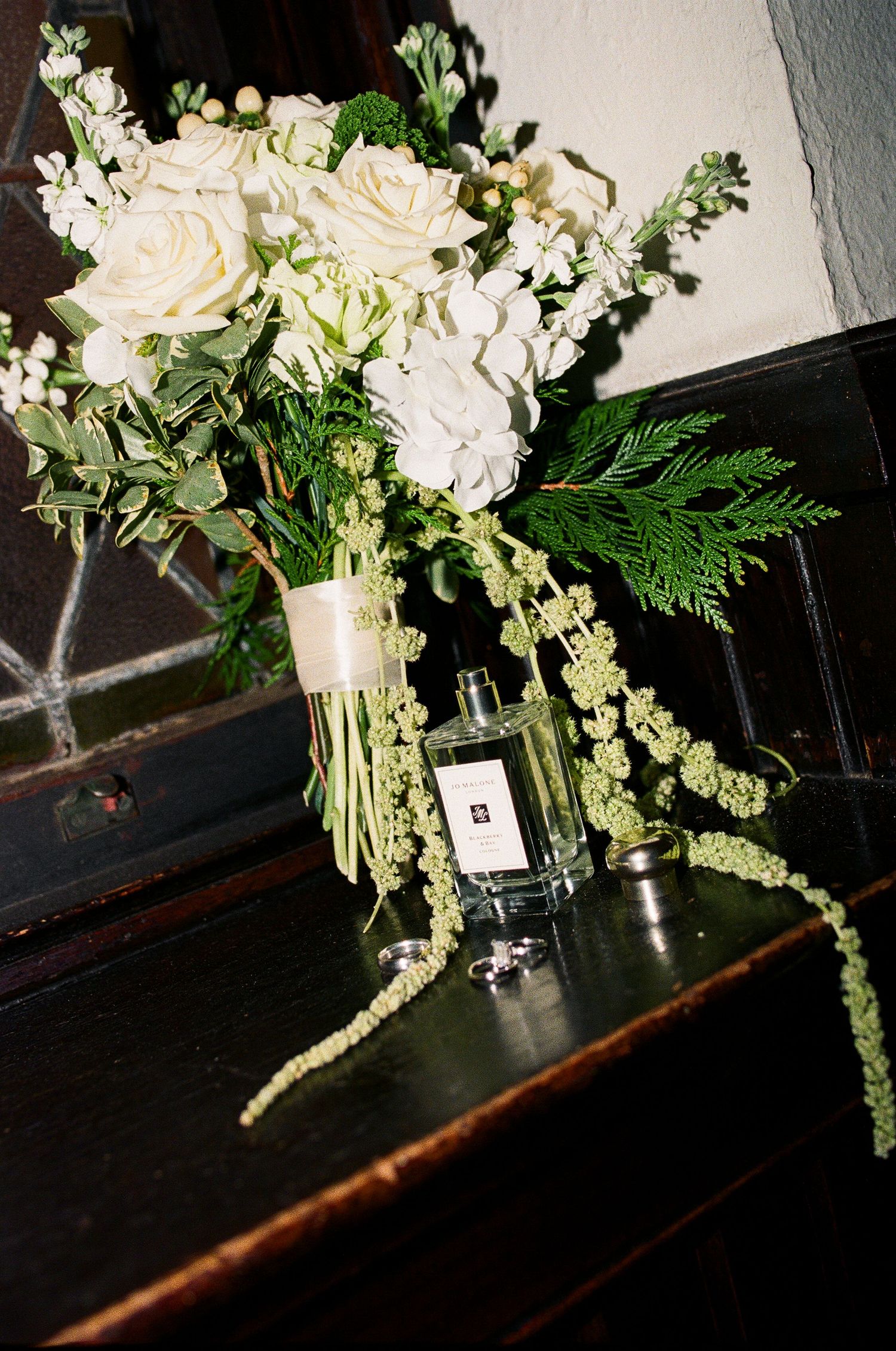 White rose wedding bouquet with greenery beside Jo Malone perfume and rings on dark wood surface.