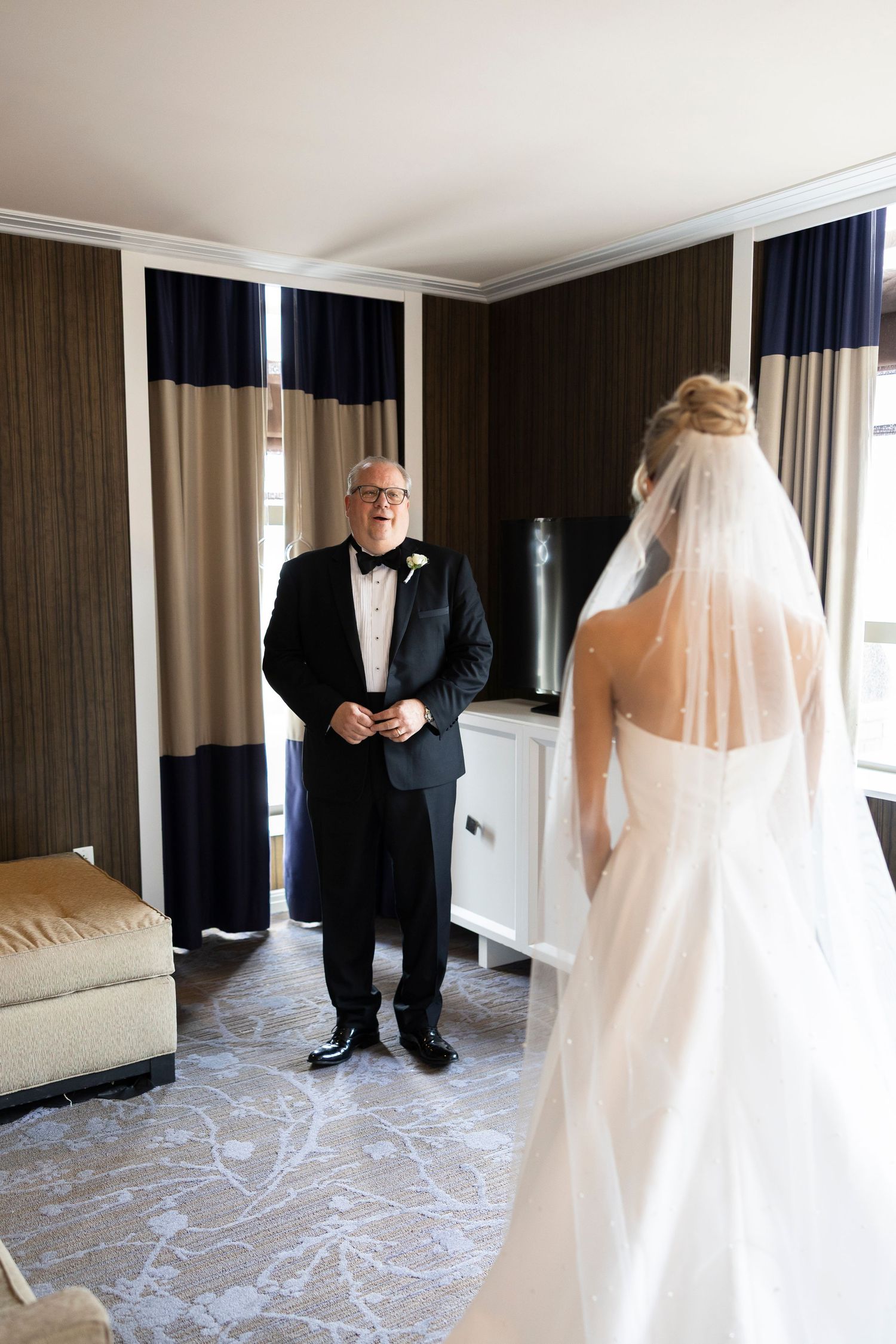 Father sees bride in wedding dress for the first time, emotional first look moment in elegant hotel room.