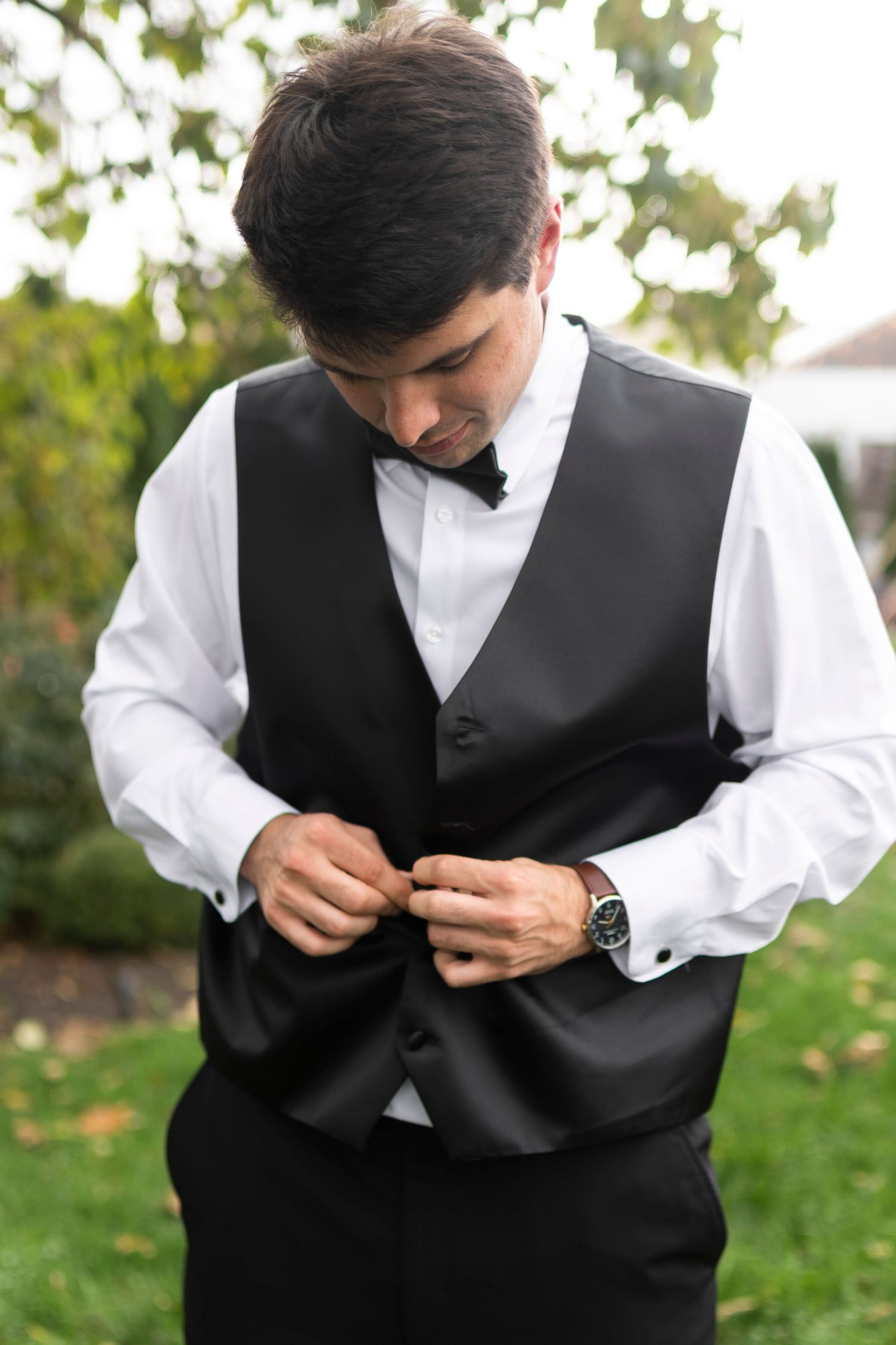 Groom in formal attire buttoning black vest outdoors.