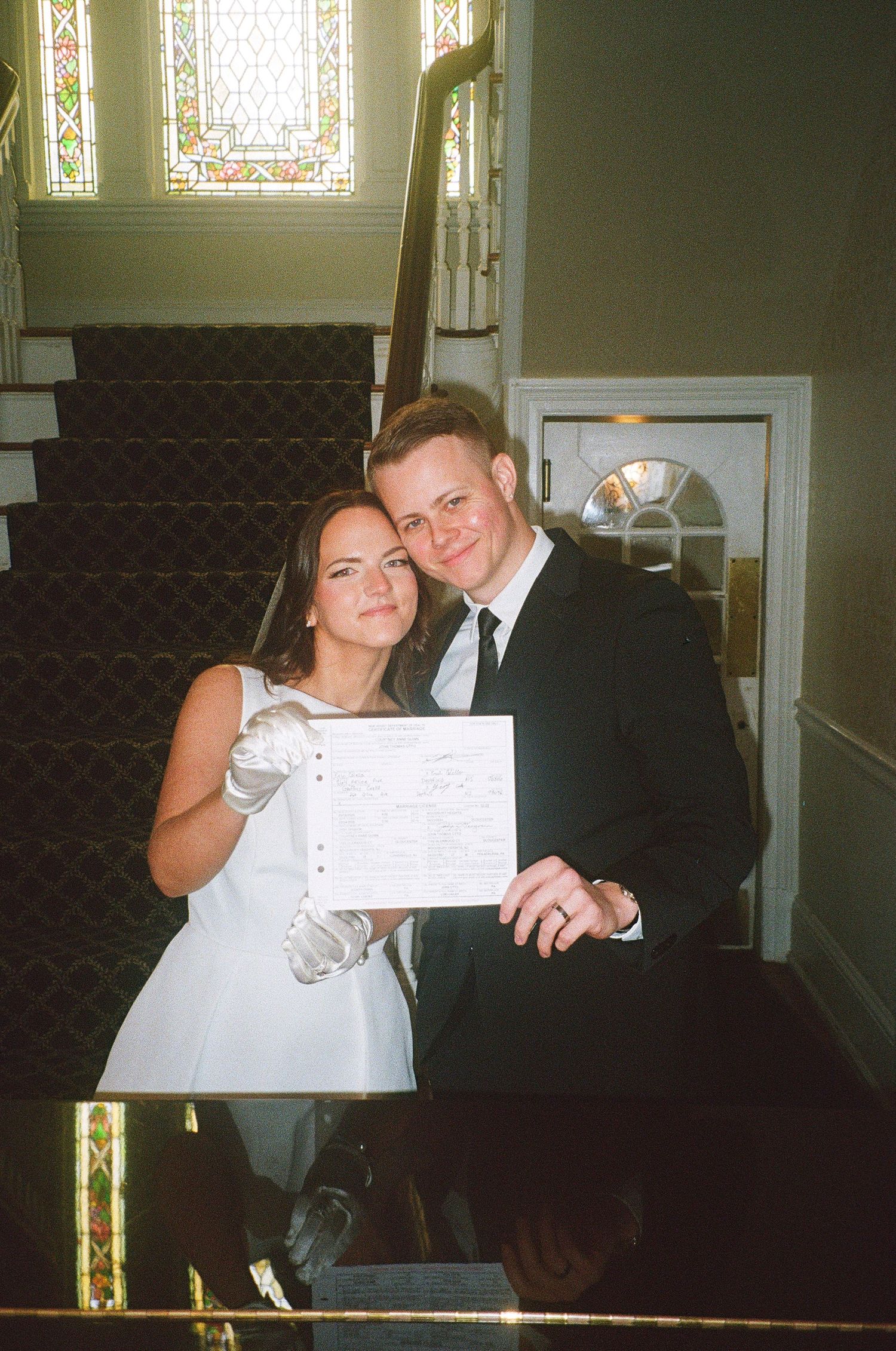A couple in wedding attire holds up paperwork while sitting in a church pew.