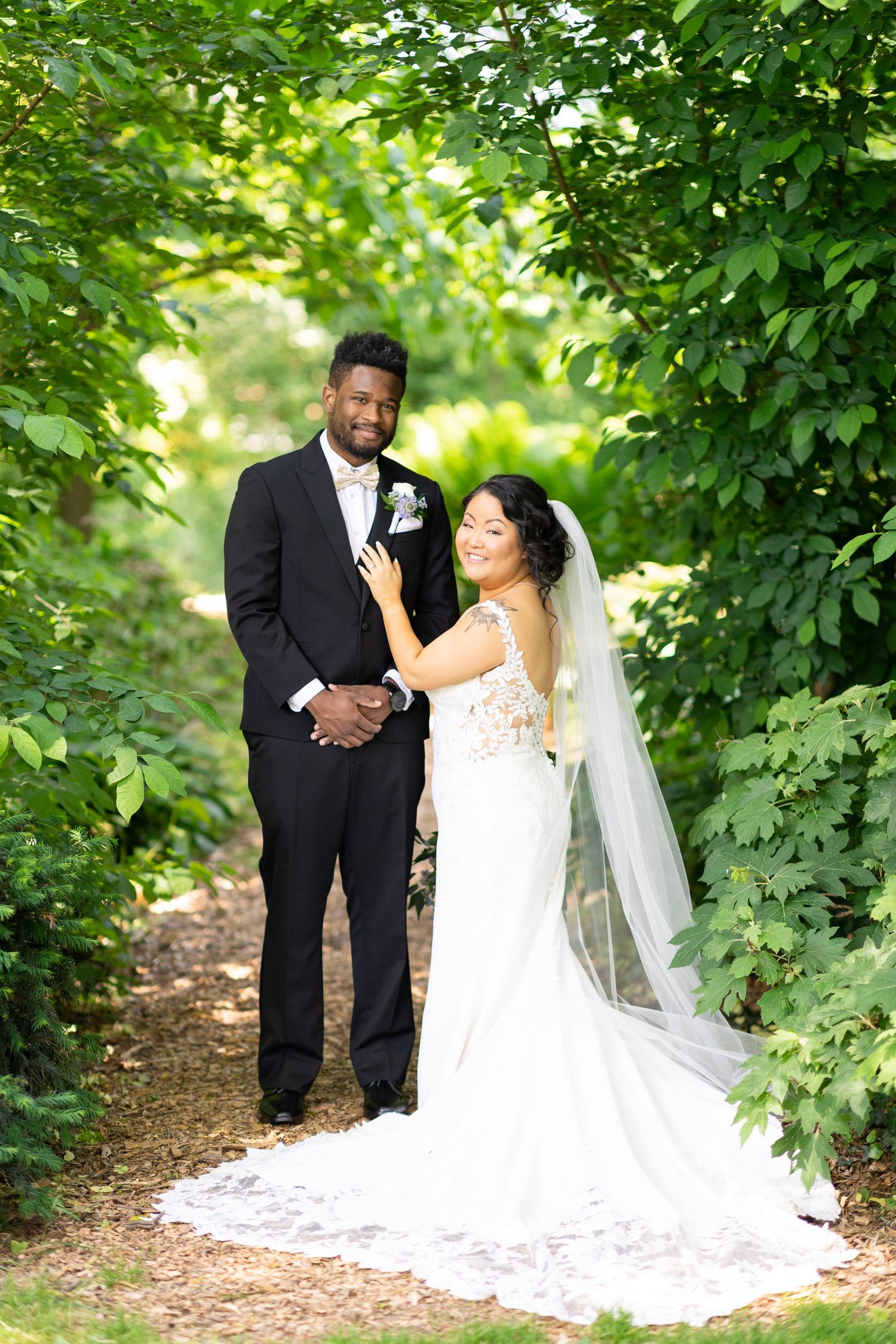 A wedding couple poses together on a wooded path with lush green foliage and natural lighting.