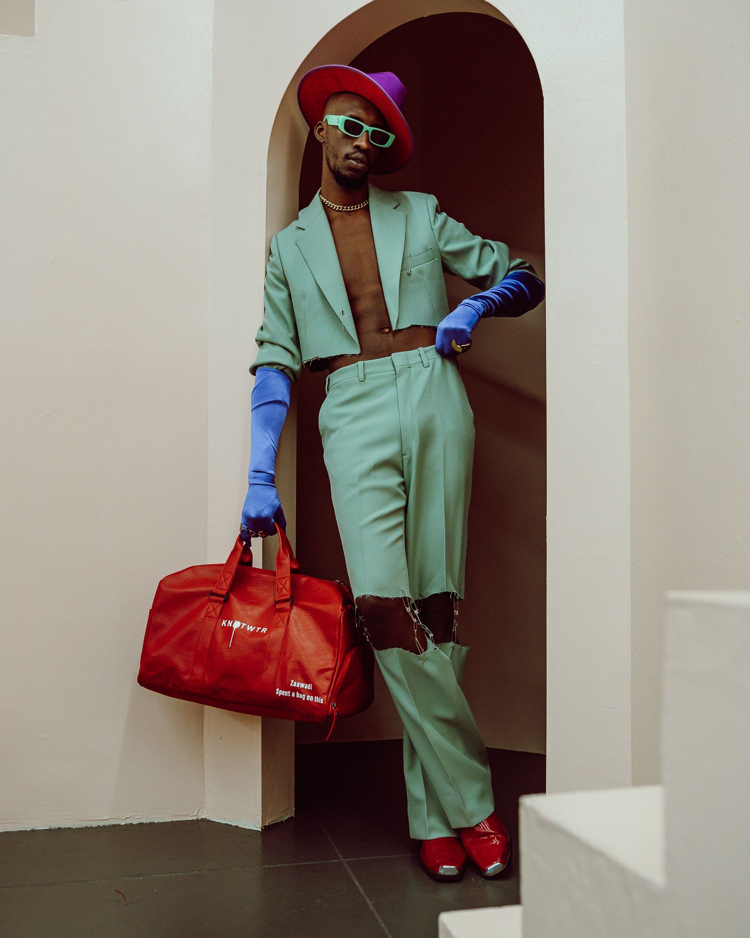 Fashion pose in mint green suit with blue gloves and red accessories against white archway. PZ STUDIO VISUALS