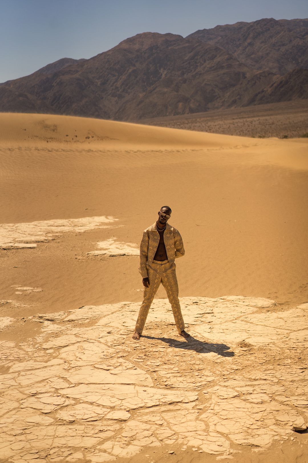A figure in formal attire poses dramatically on cracked desert ground with mountains in the background. PZ STUDIO VISUALS