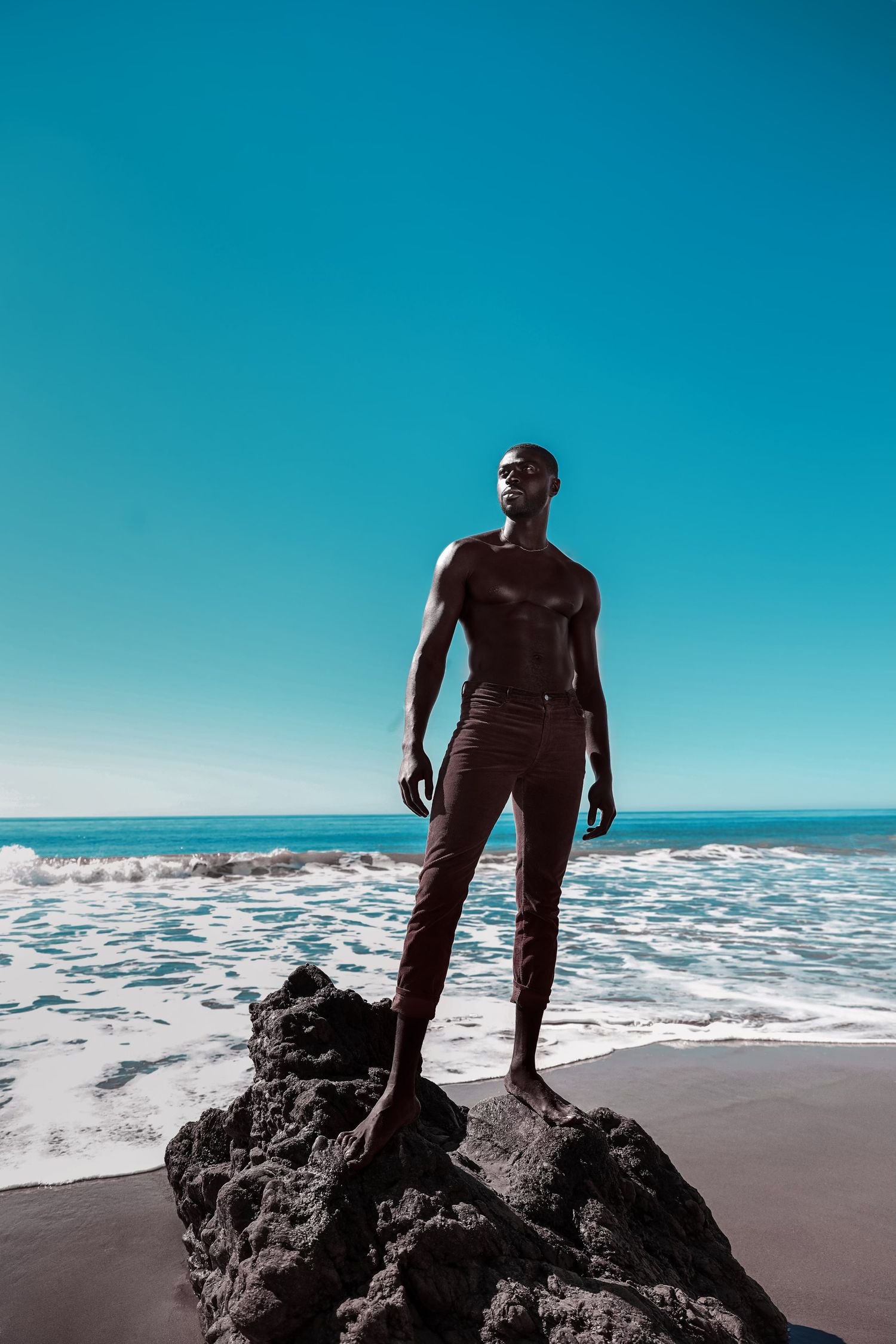 Silhouetted figure explores rocky beach landscape against bright blue sky and ocean waves. Julius Antwi for PZ STUDIO VISUALS