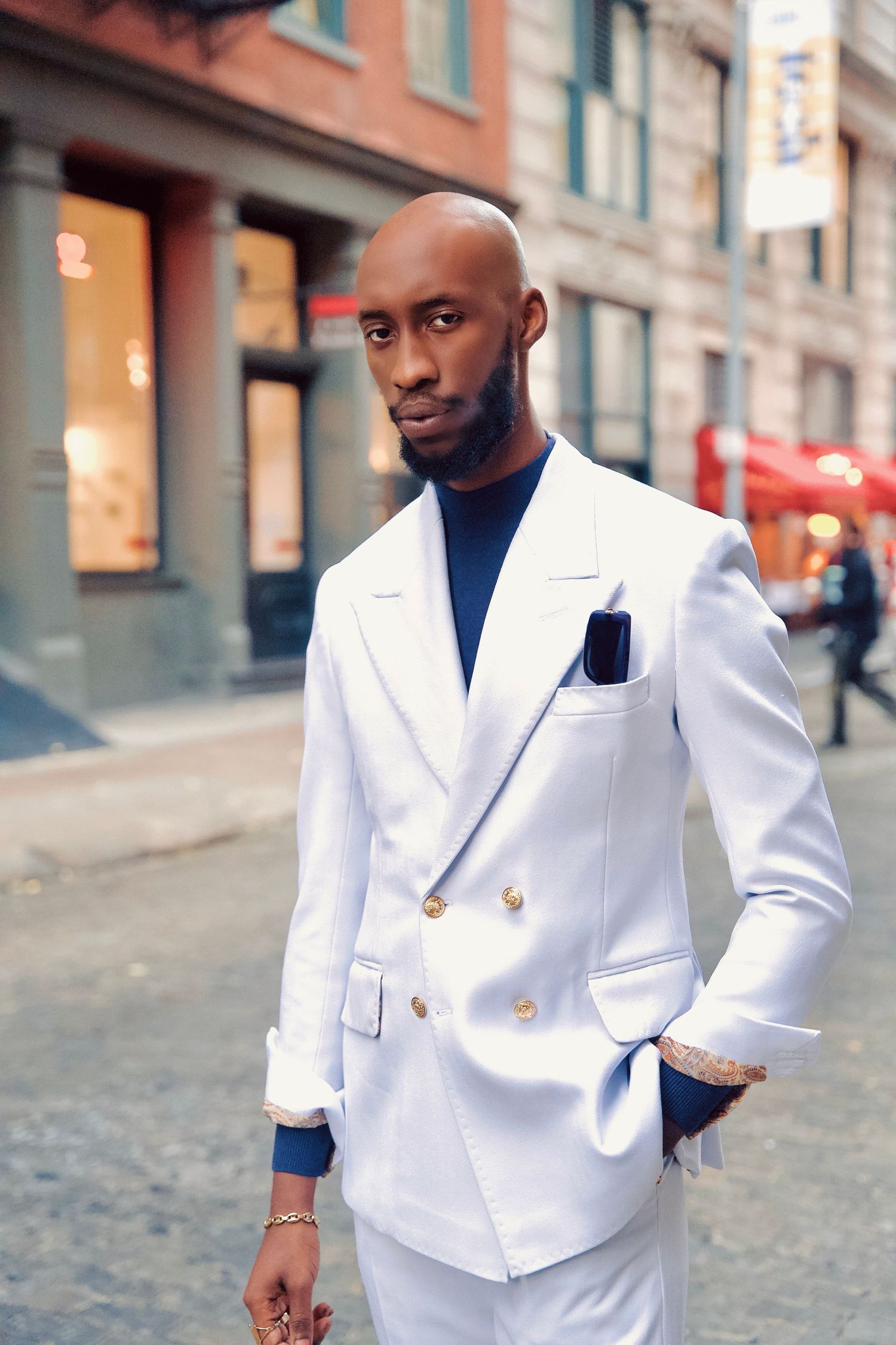 A person wearing a crisp white blazer poses on a cobblestone street in an urban setting.
