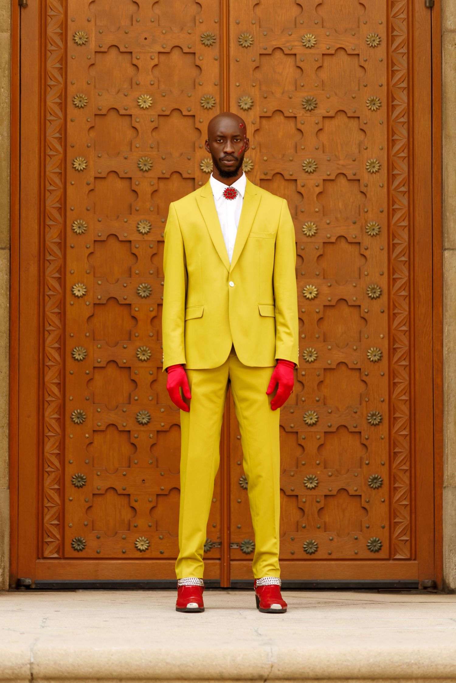 Figure in bright yellow suit and red gloves poses against ornate wooden door with geometric patterns. PZ STUDIO VISUALS  x Beoncia Dunn