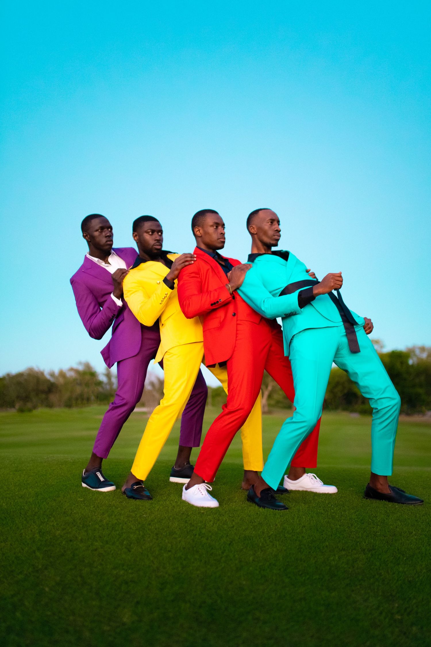 Performers in colorful suits create domino effect pose on green grass against blue sky. PZ STUDIO VISUALS