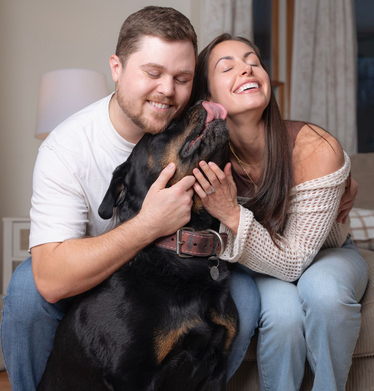Couple snuggling with their black Rottweiler dog on a light colored couch.