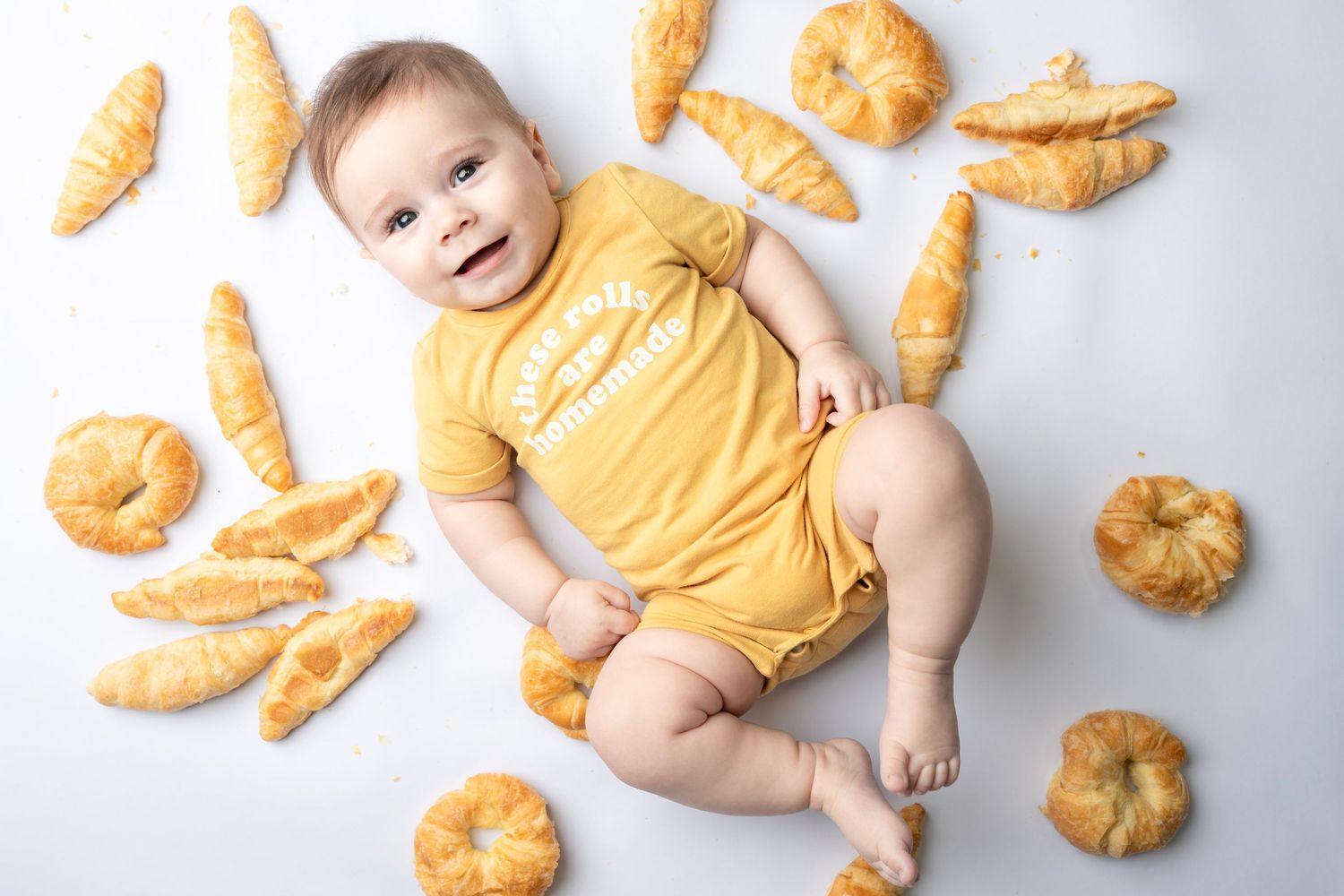 Baby in yellow onesie lying on white background surrounded by croissants and pastries.