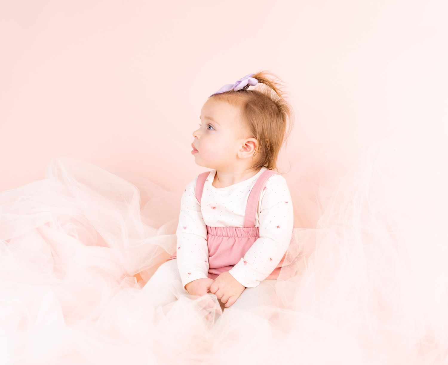 Baby in pink dress poses against soft pink background in dreamy portrait series.