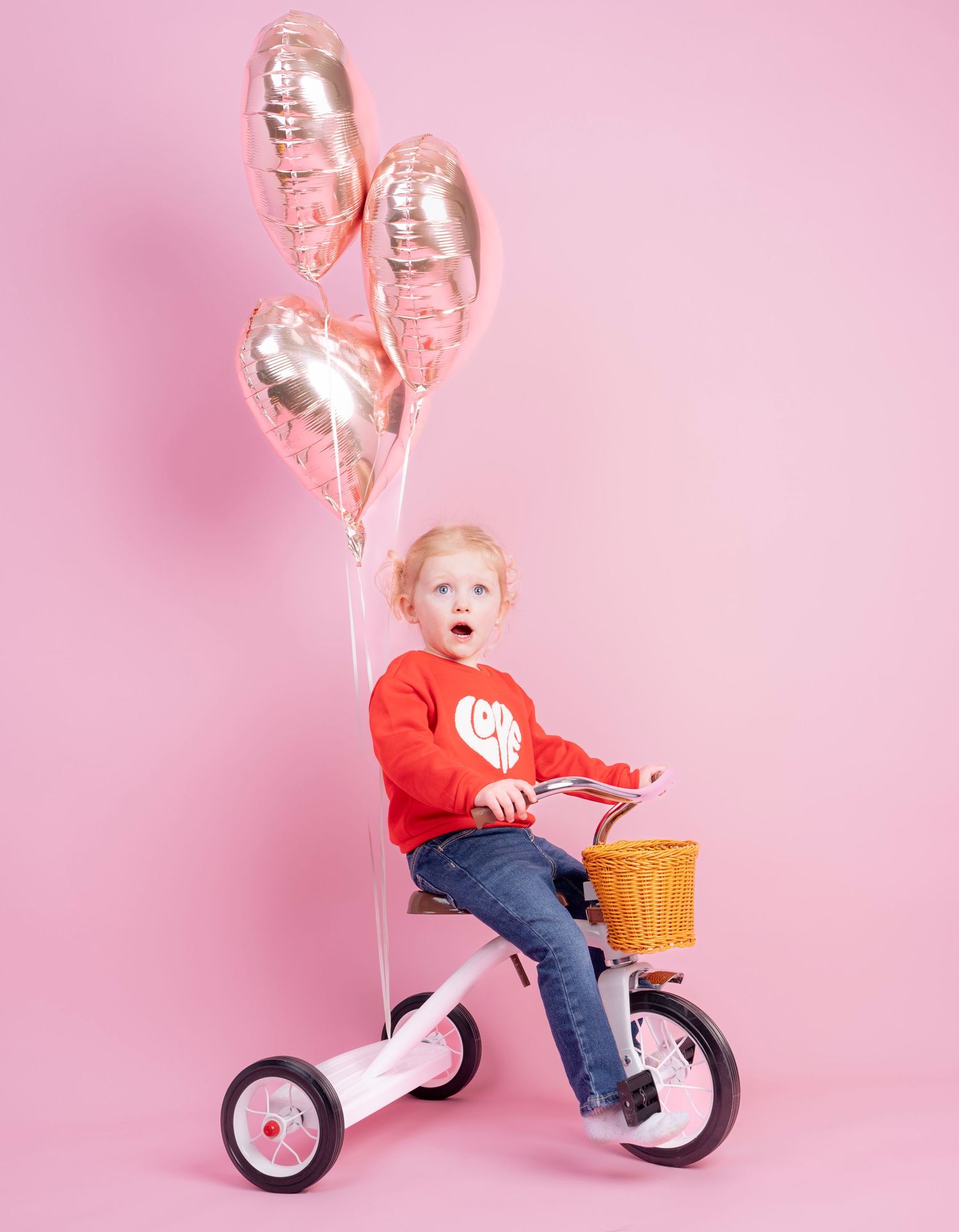 Child riding a red tricycle with pink balloons against pink background.