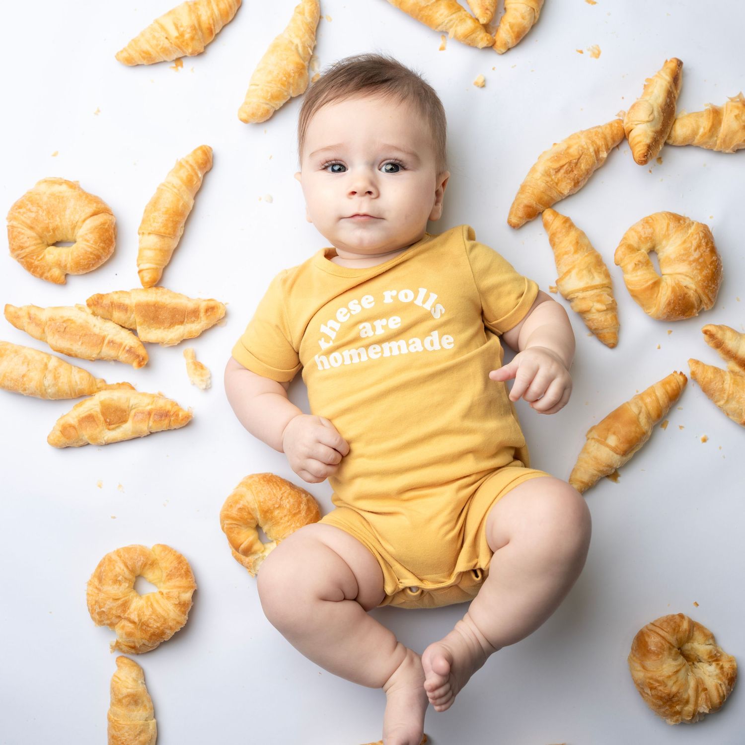 Adorable baby in yellow onesie lying on white background surrounded by croissant-shaped pastries.