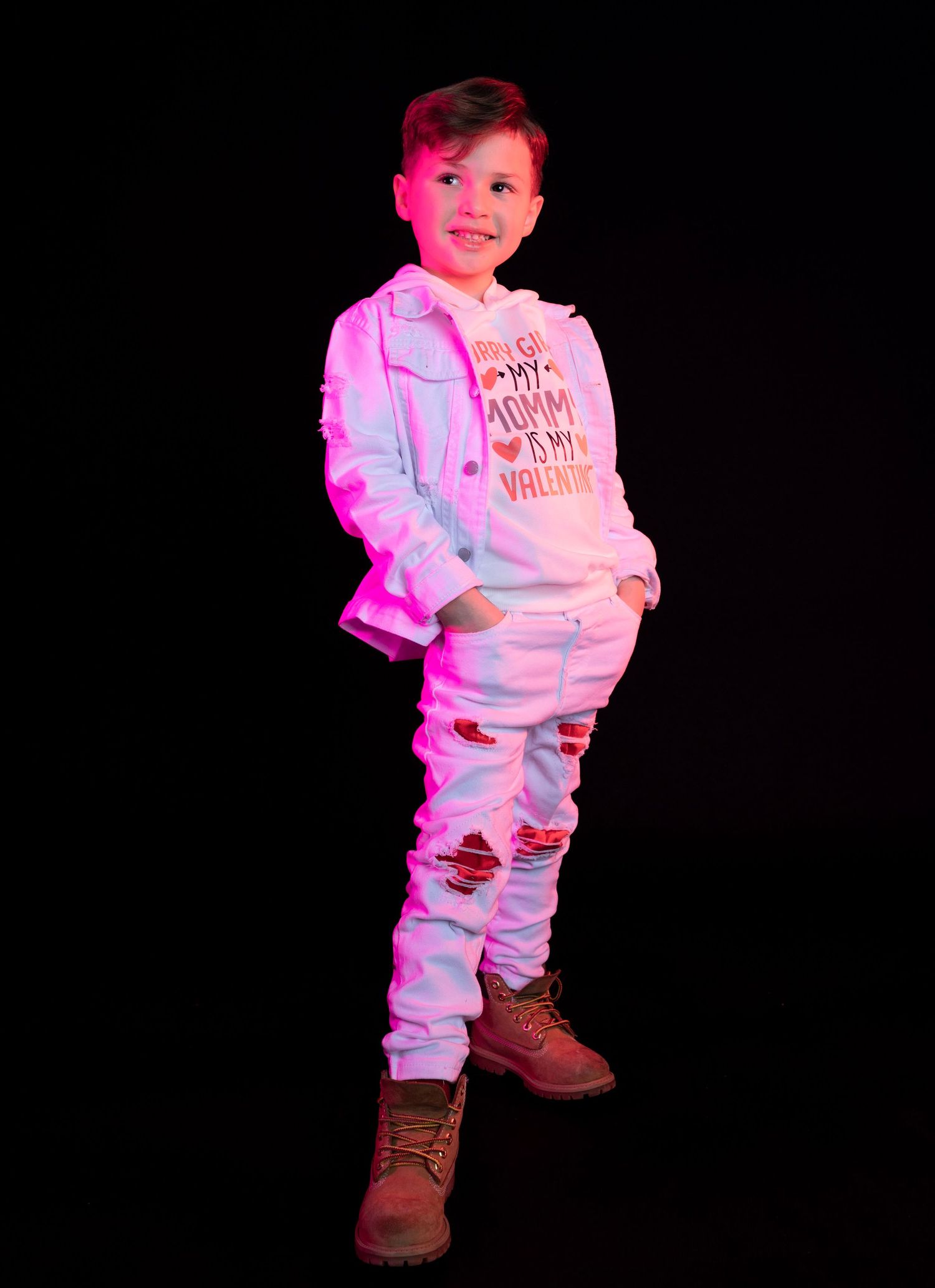Young child in white outfit poses against dark background with pink lighting effects.