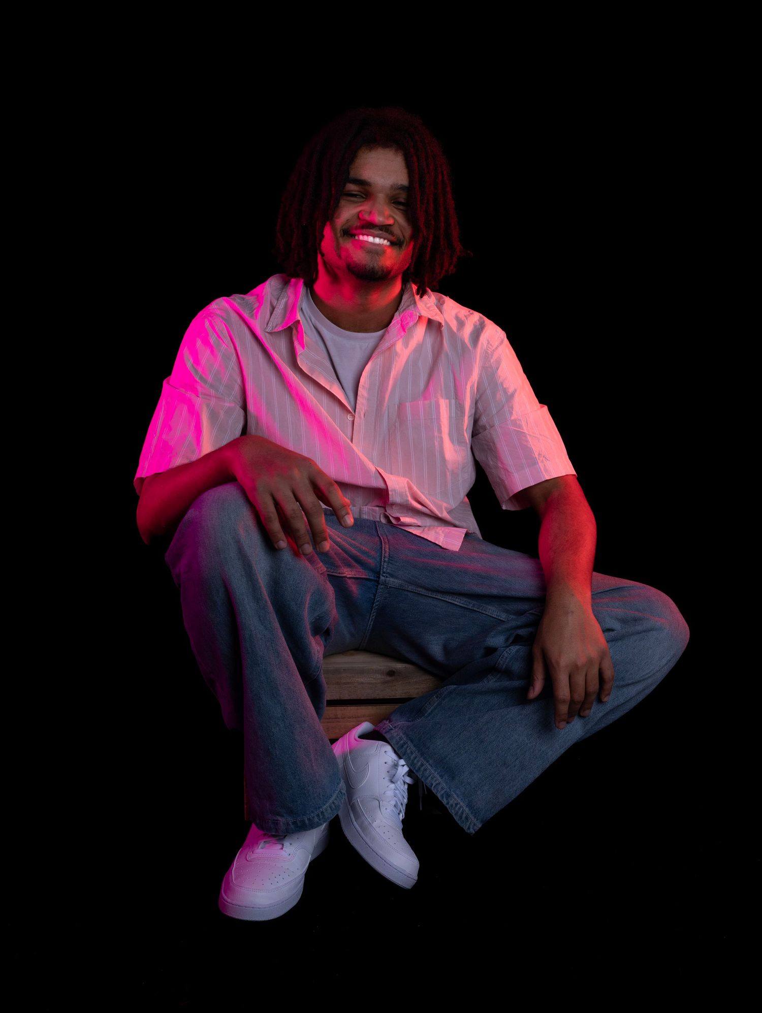 Person in casual attire sitting against dark background with dramatic pink and blue lighting.