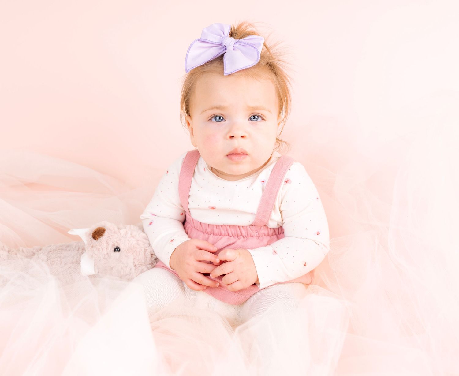 Child in pink dress with bow sitting on light pink background.
