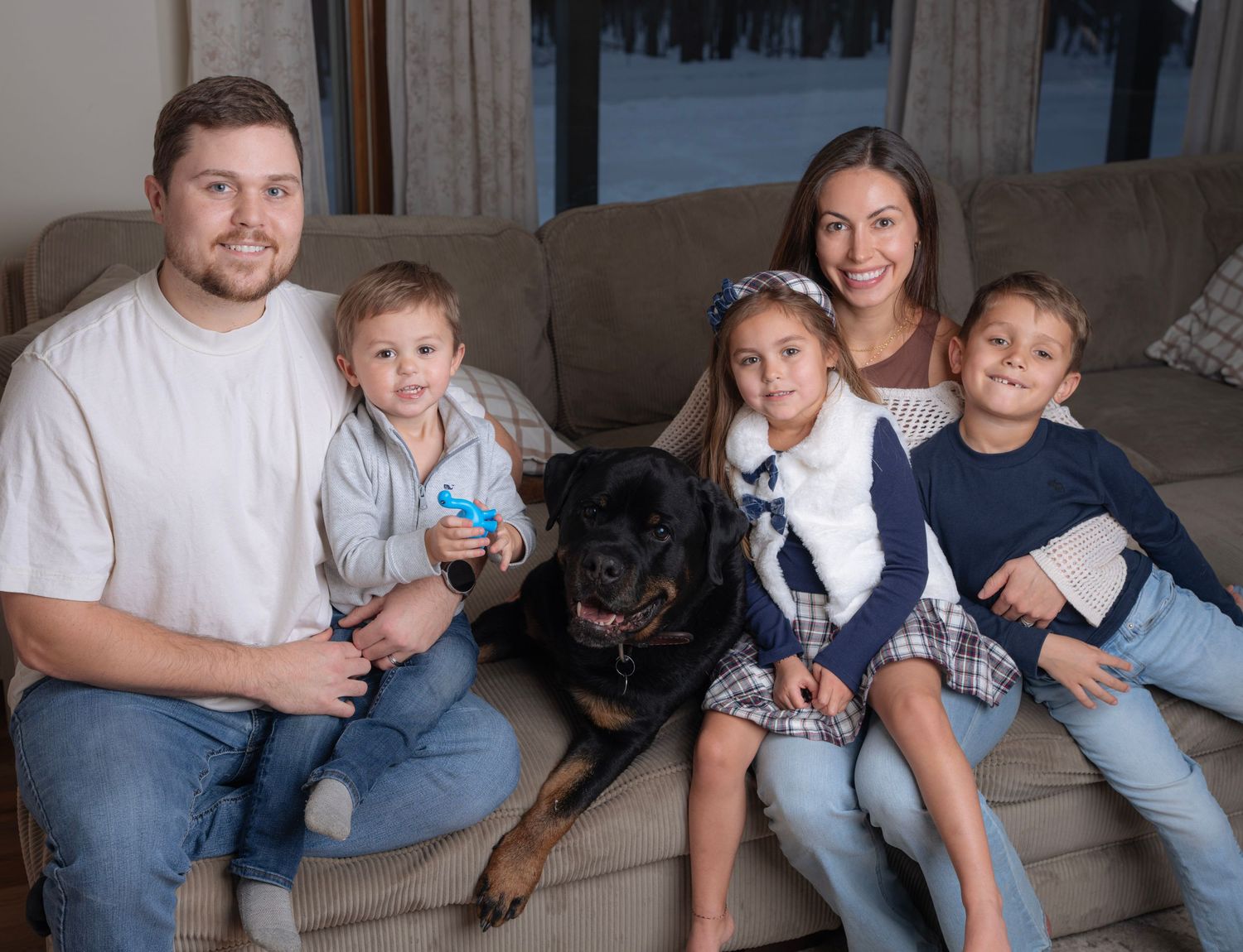 A family sits together on a couch with their black Rottweiler dog in a casual indoor setting.