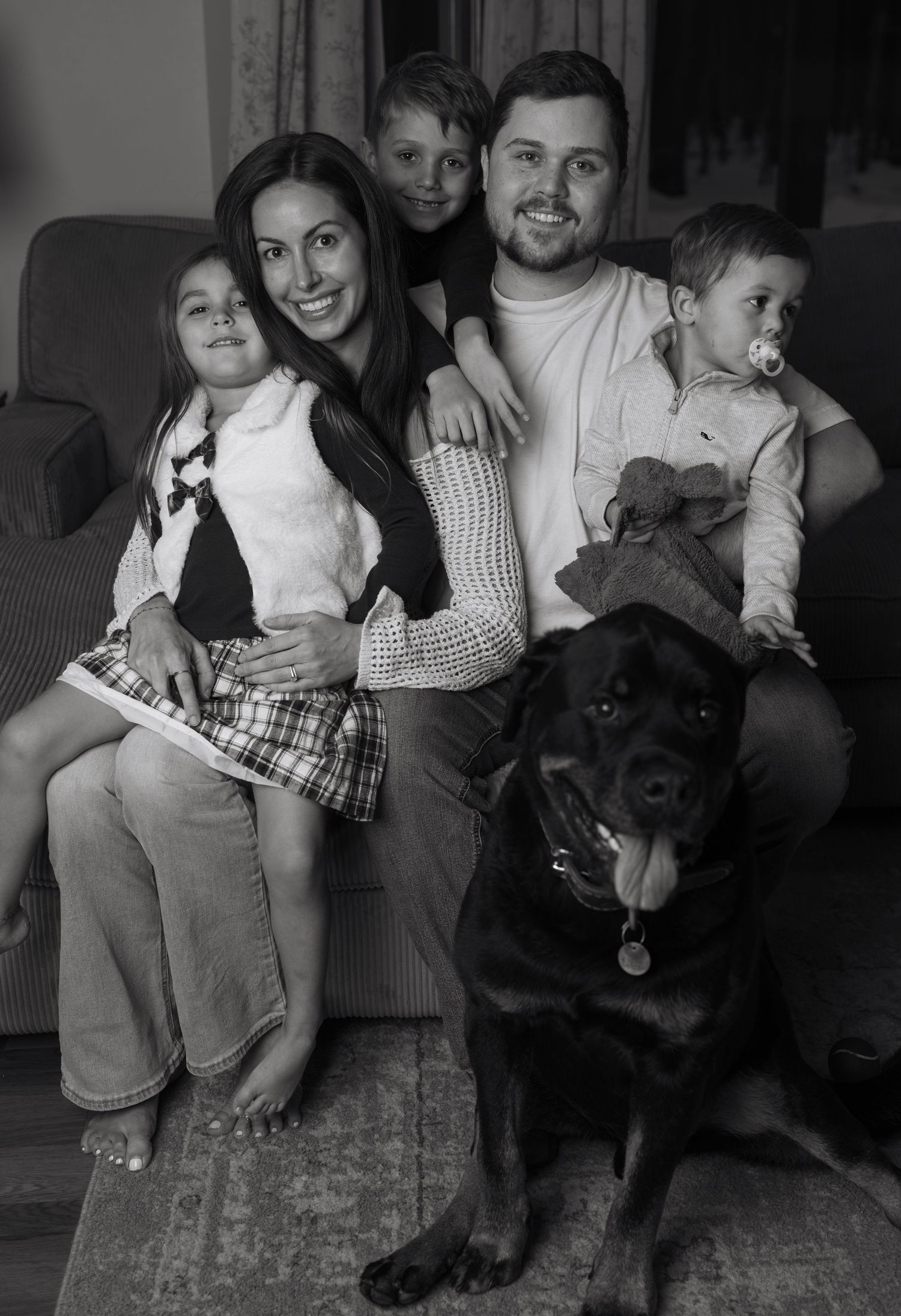 Family portrait on a couch with a black Rottweiler dog in casual indoor setting.