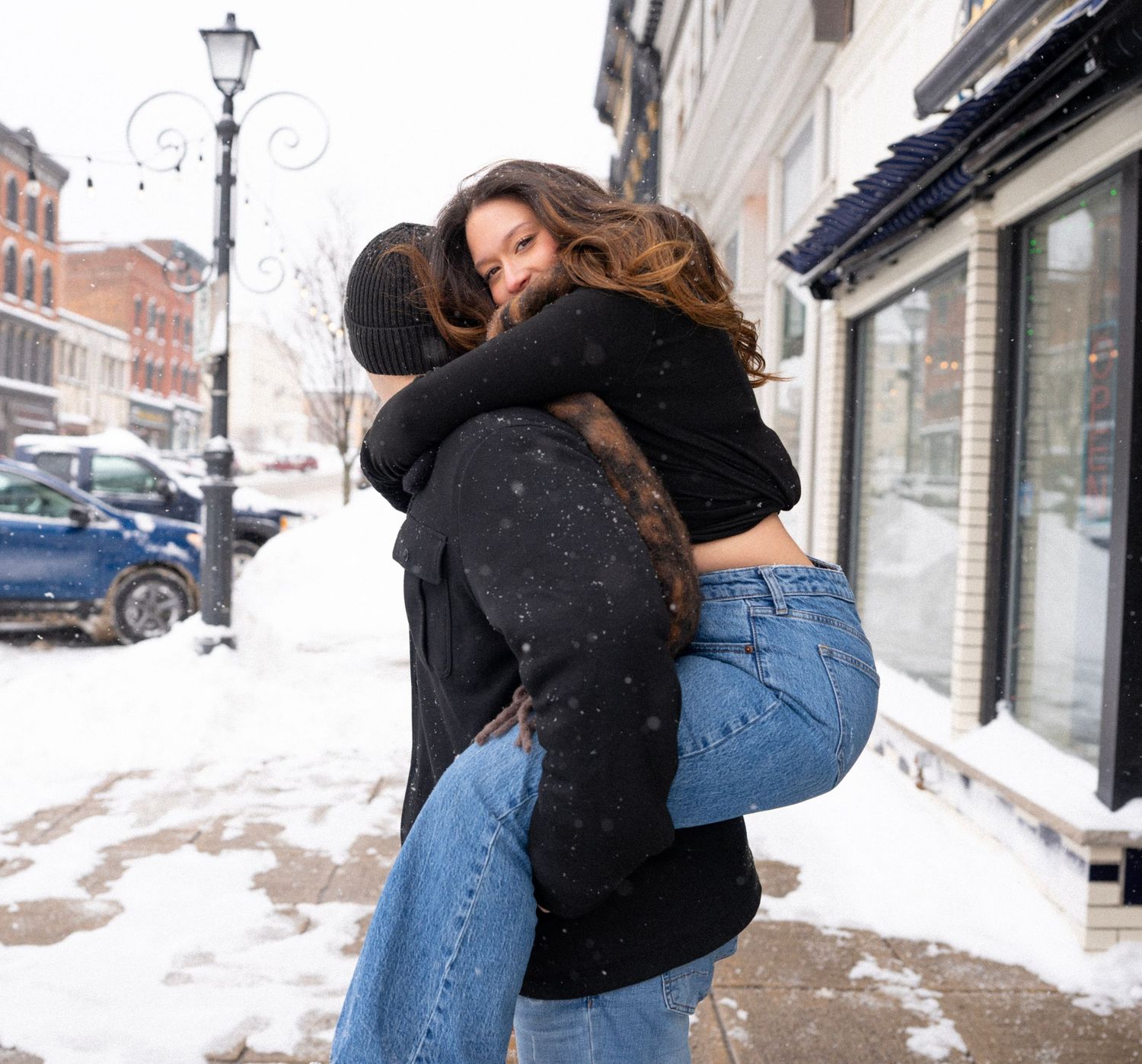 A playful winter embrace between a couple on a snowy city sidewalk.