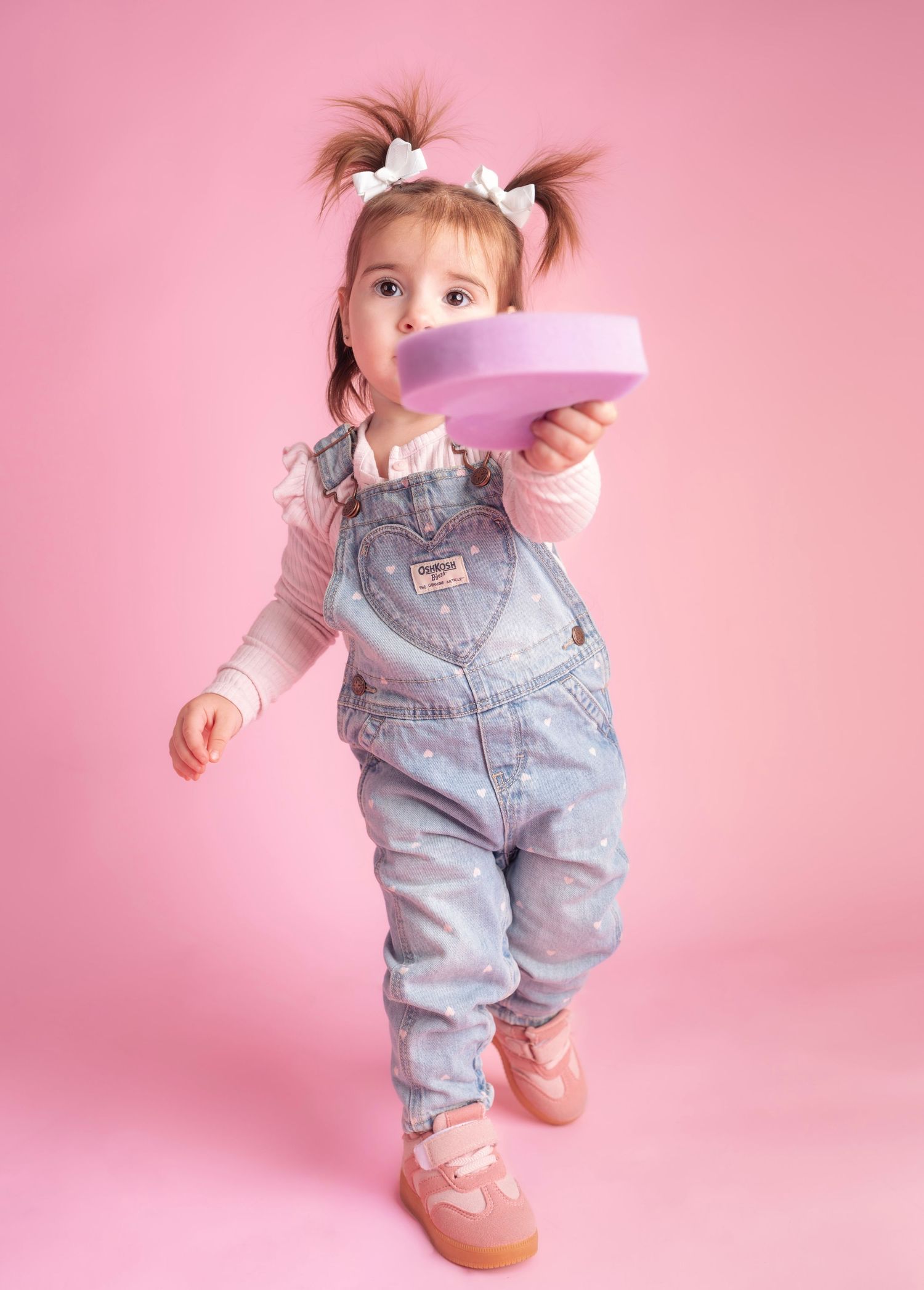 Small child wearing denim overalls stands against pink background in playful poses.