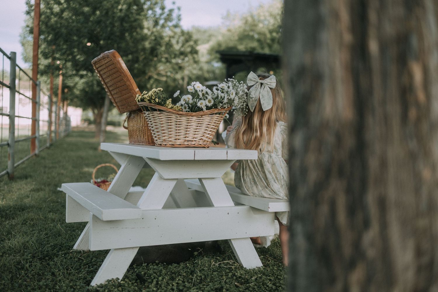 A wicker basket with flowers sits on a small white picnic table in a green outdoor setting.