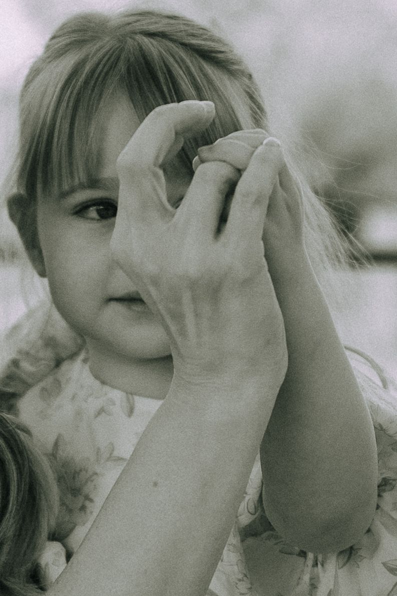 Black and white close-up of a young girl peering through an adults hands forming a heart shape.