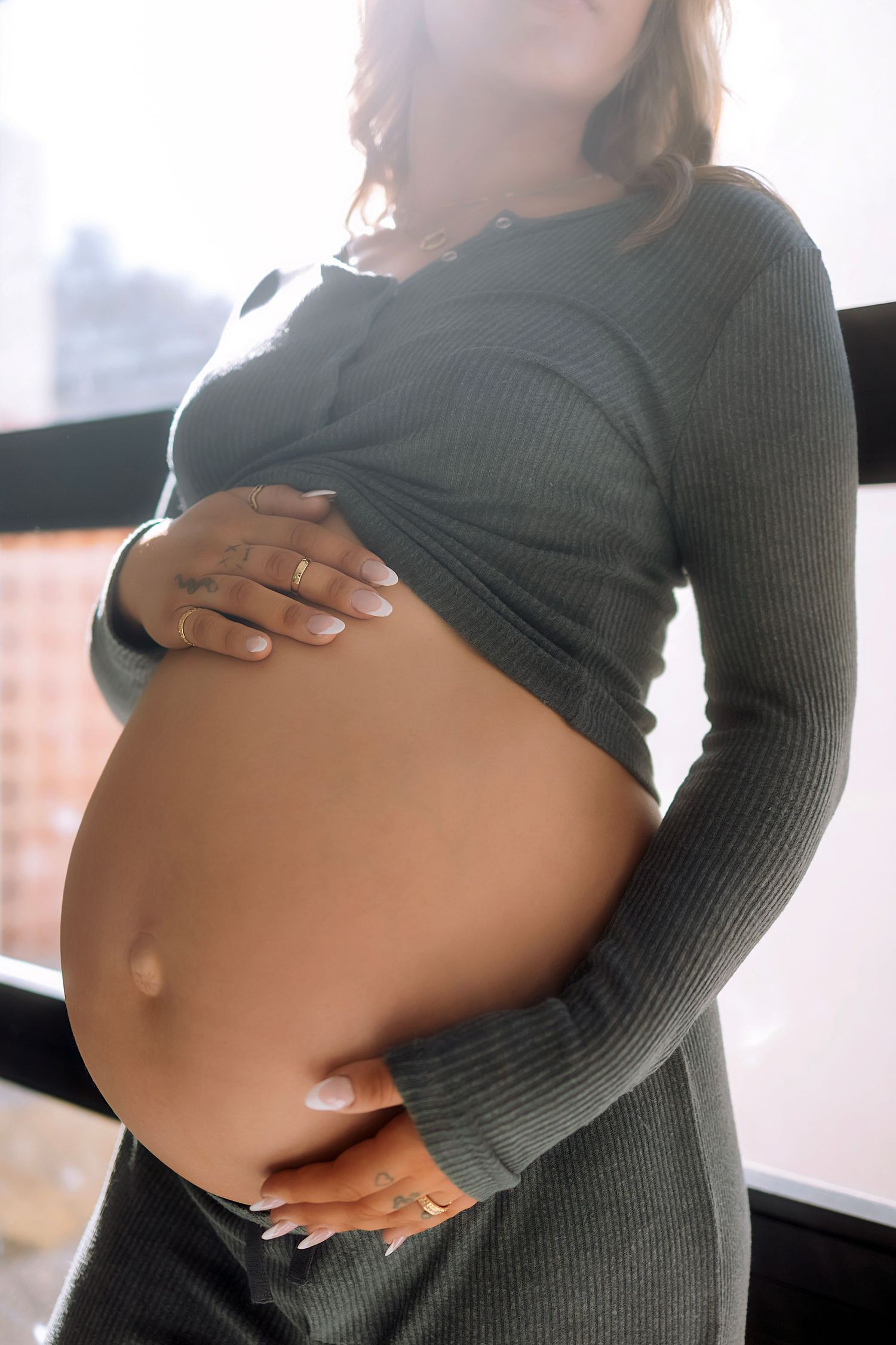 Pregnant woman in gray ribbed top cradling baby bump by window.