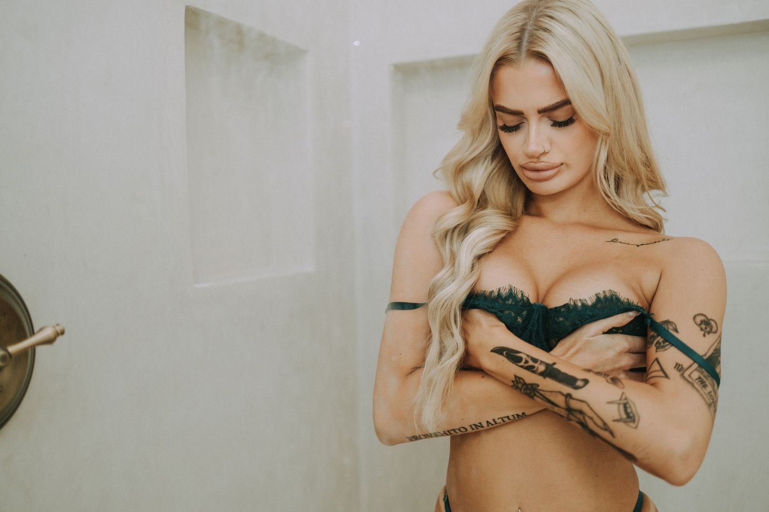 Blonde tattooed woman in green lace bralette posing in bright bathroom.