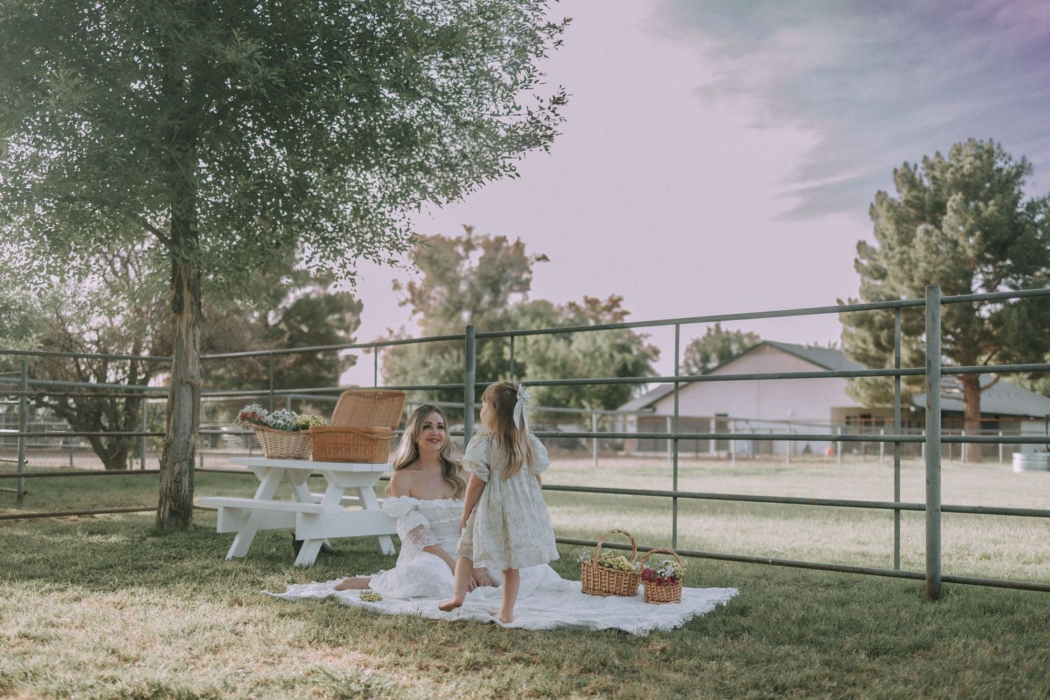 A woman and young girl in white dresses sit together on a picnic blanket under a tree.