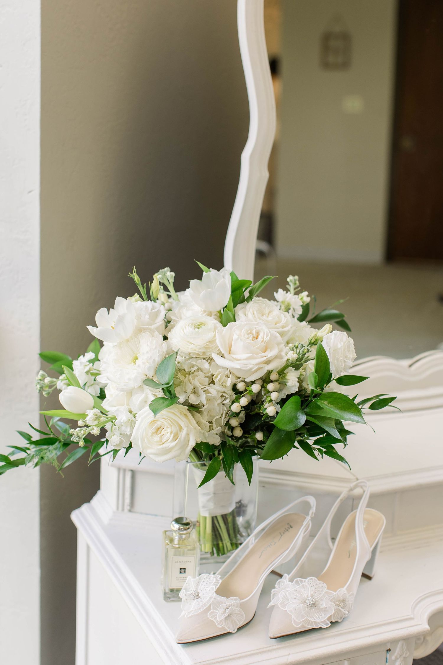 White roses and bridal shoes arranged on white table with greenery accents.