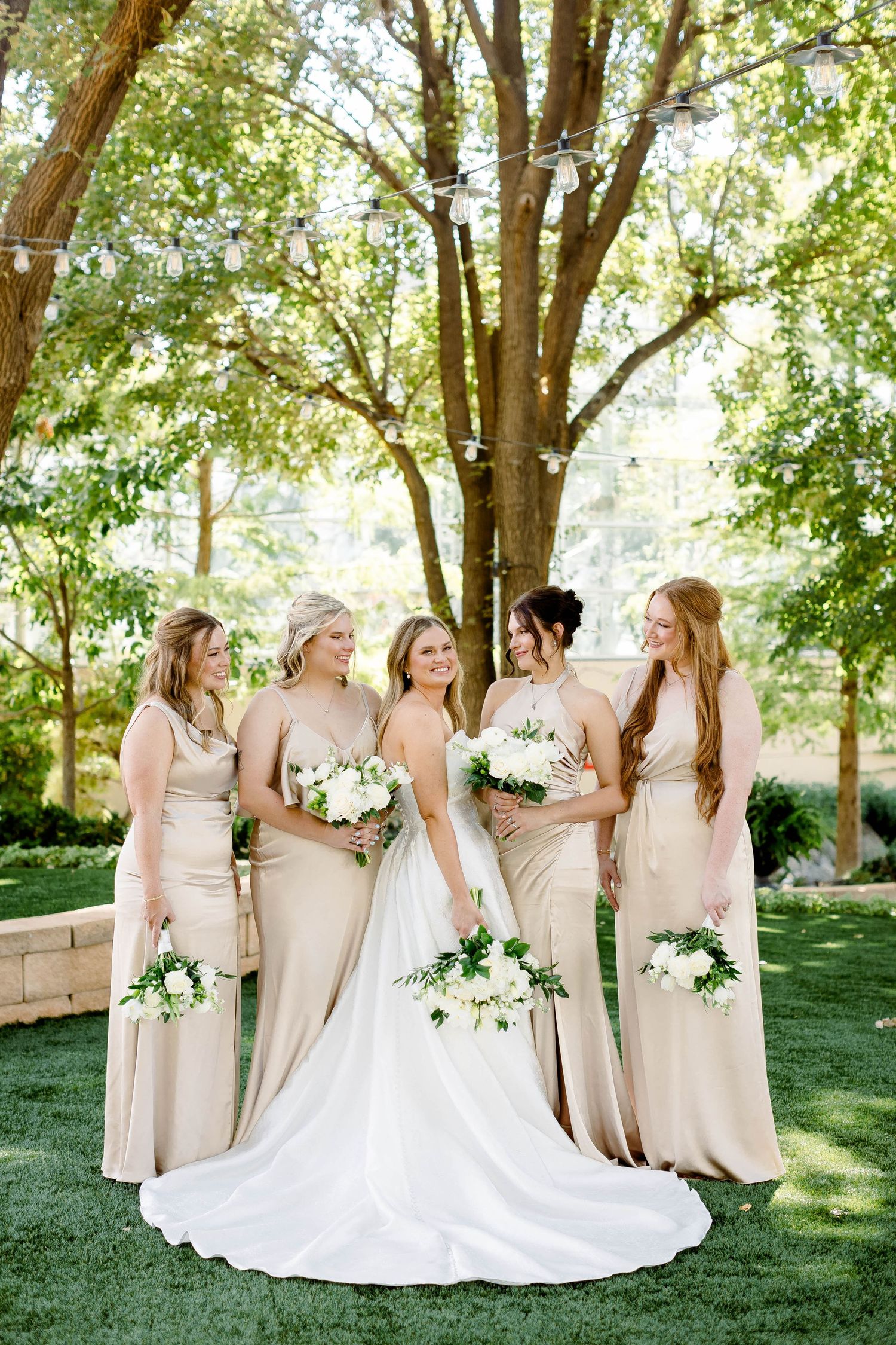 Wedding party arranged in elegant poses on grass with trees creating a natural backdrop.
