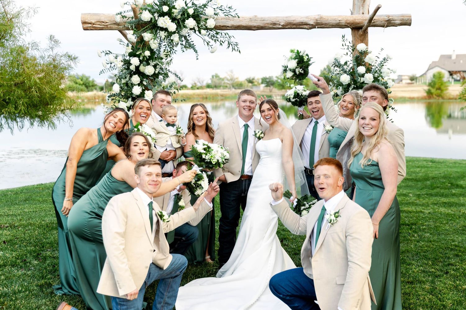 Wedding party in green dresses and tan suits poses together by a lakeside decorated with white floral arrangements.