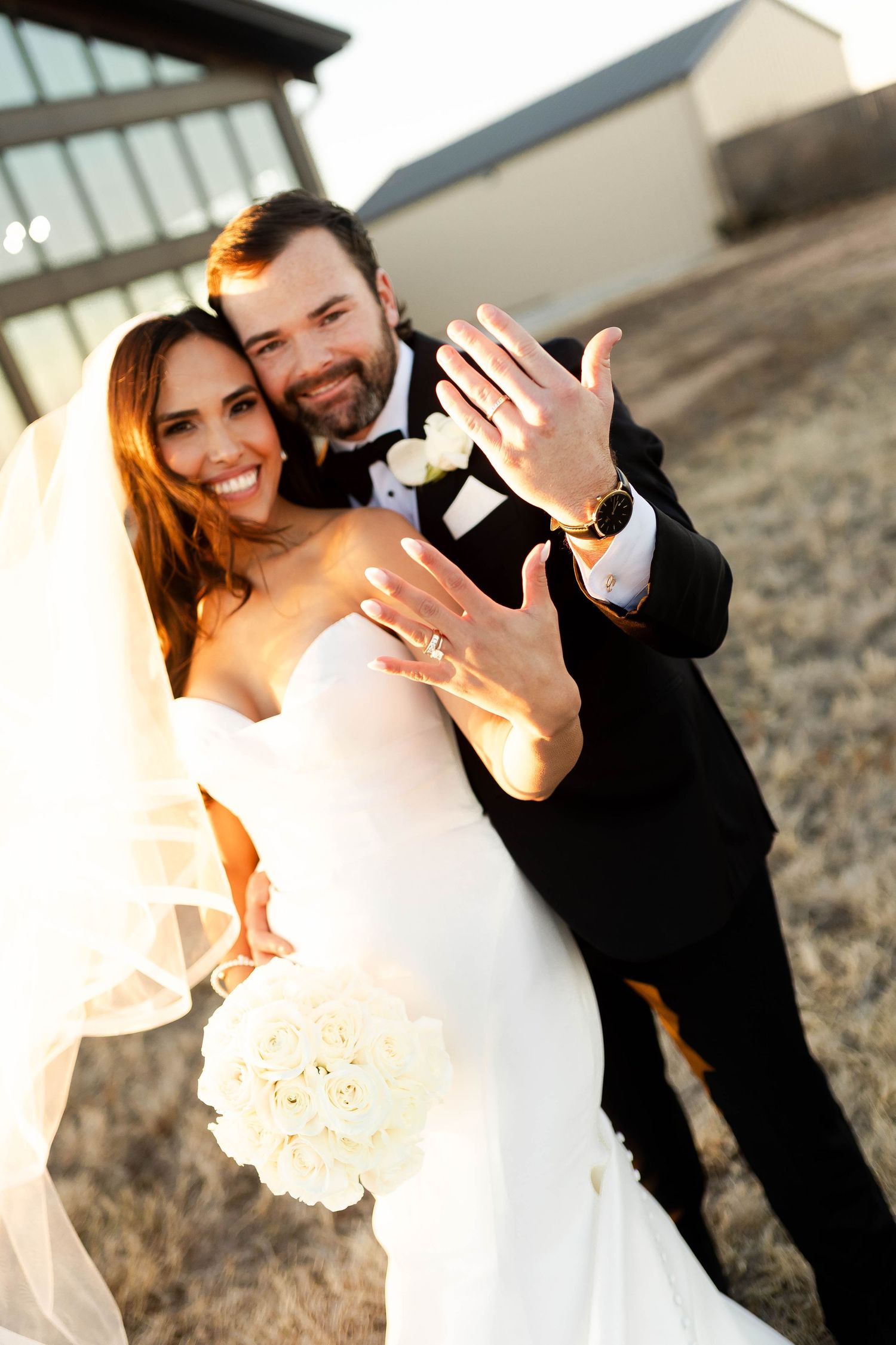 A joyful wedding couple shows off their rings in warm sunset lighting.