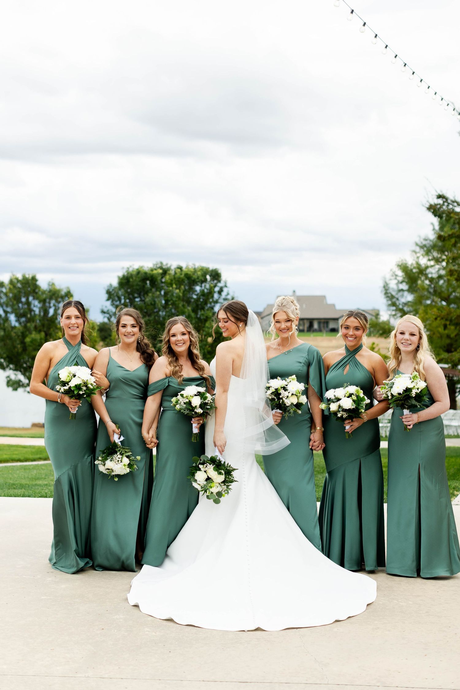 Bridesmaids in sage green dresses pose together with bride in white gown holding bouquets at outdoor wedding venue.