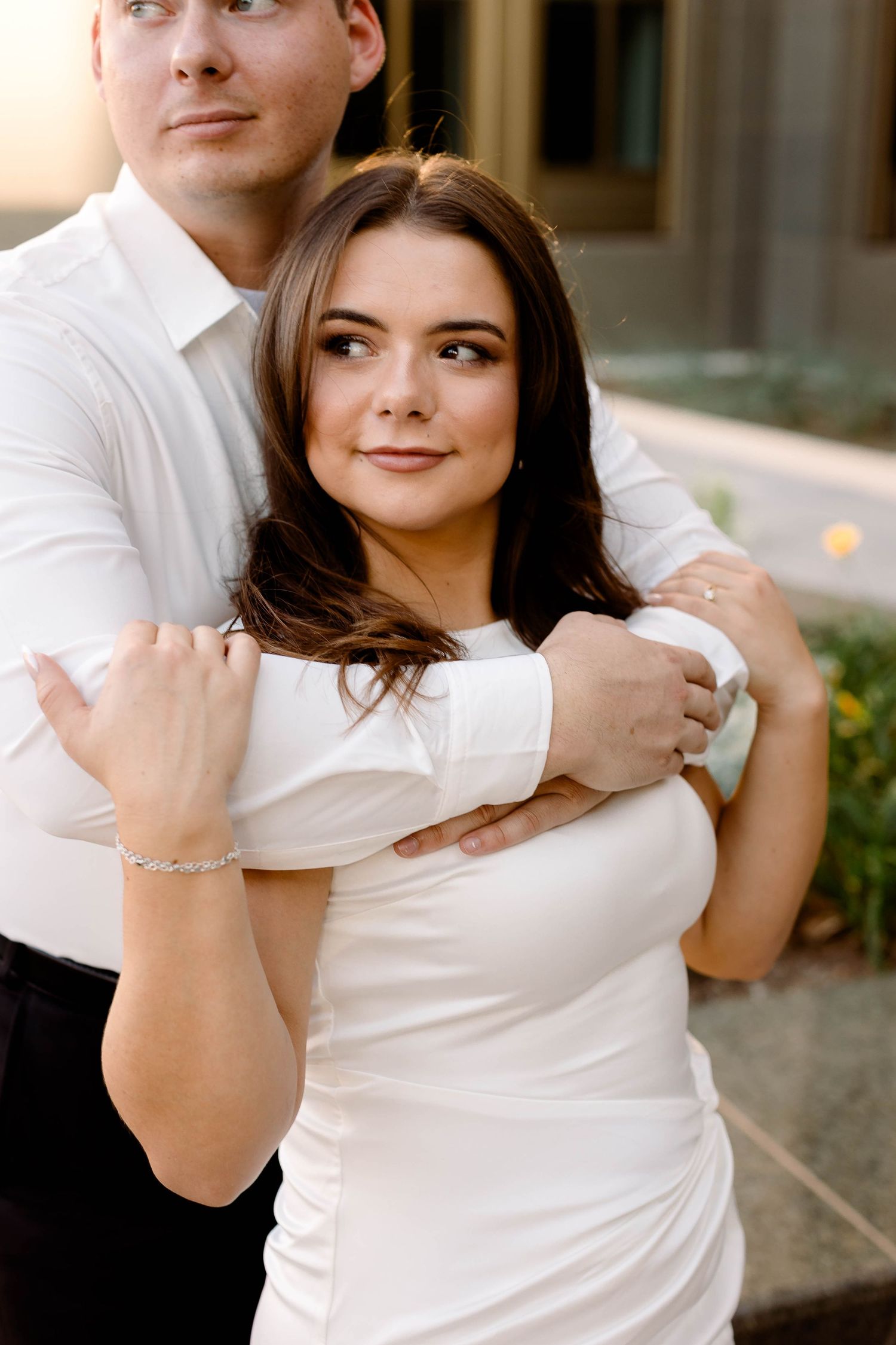 Romantic couple in white outfits sharing intimate moment against modern architecture backdrop.