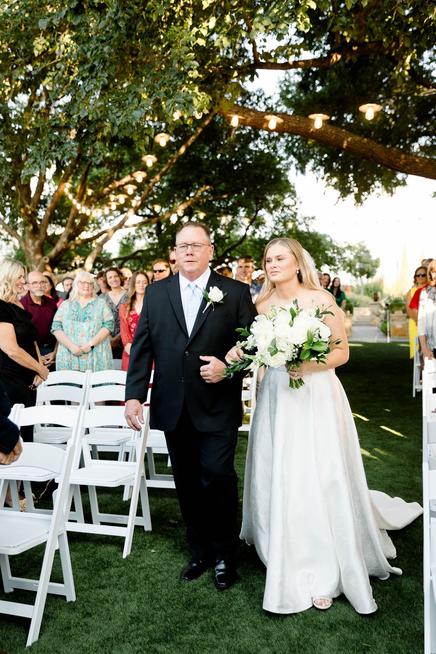 Wedding ceremony guests mingling among white folding chairs on a lawn under large shade trees.
