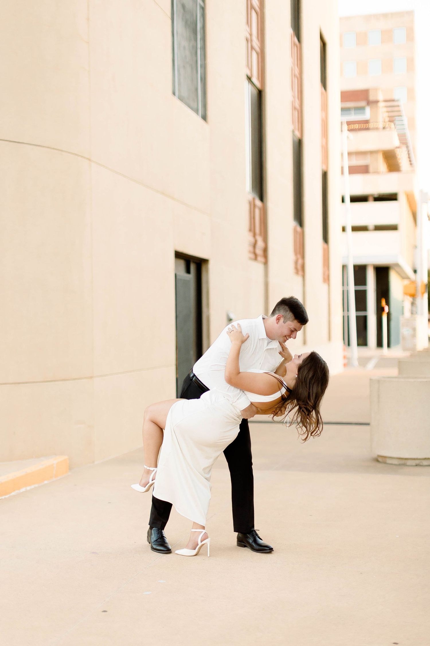 A couple poses playfully in formal wear against a beige wall outdoors.