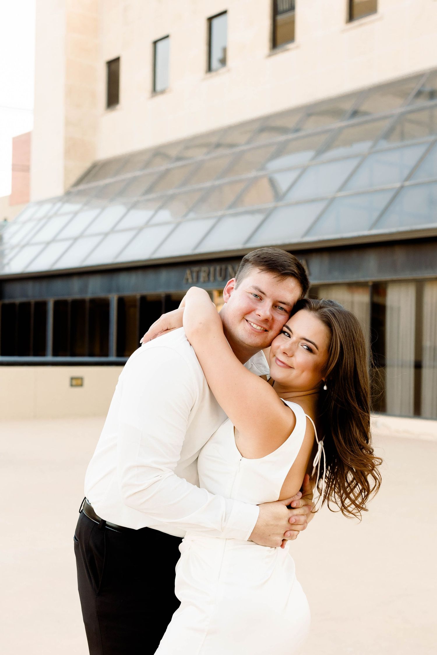 A romantic couple in white and black attire share an intimate moment against a beige building exterior.