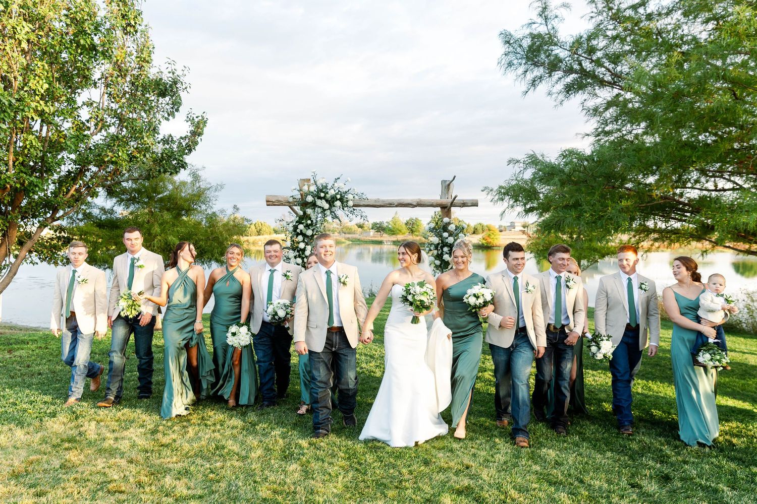 Wedding party members in sage green dresses and dark suits pose together on a grassy lawn.