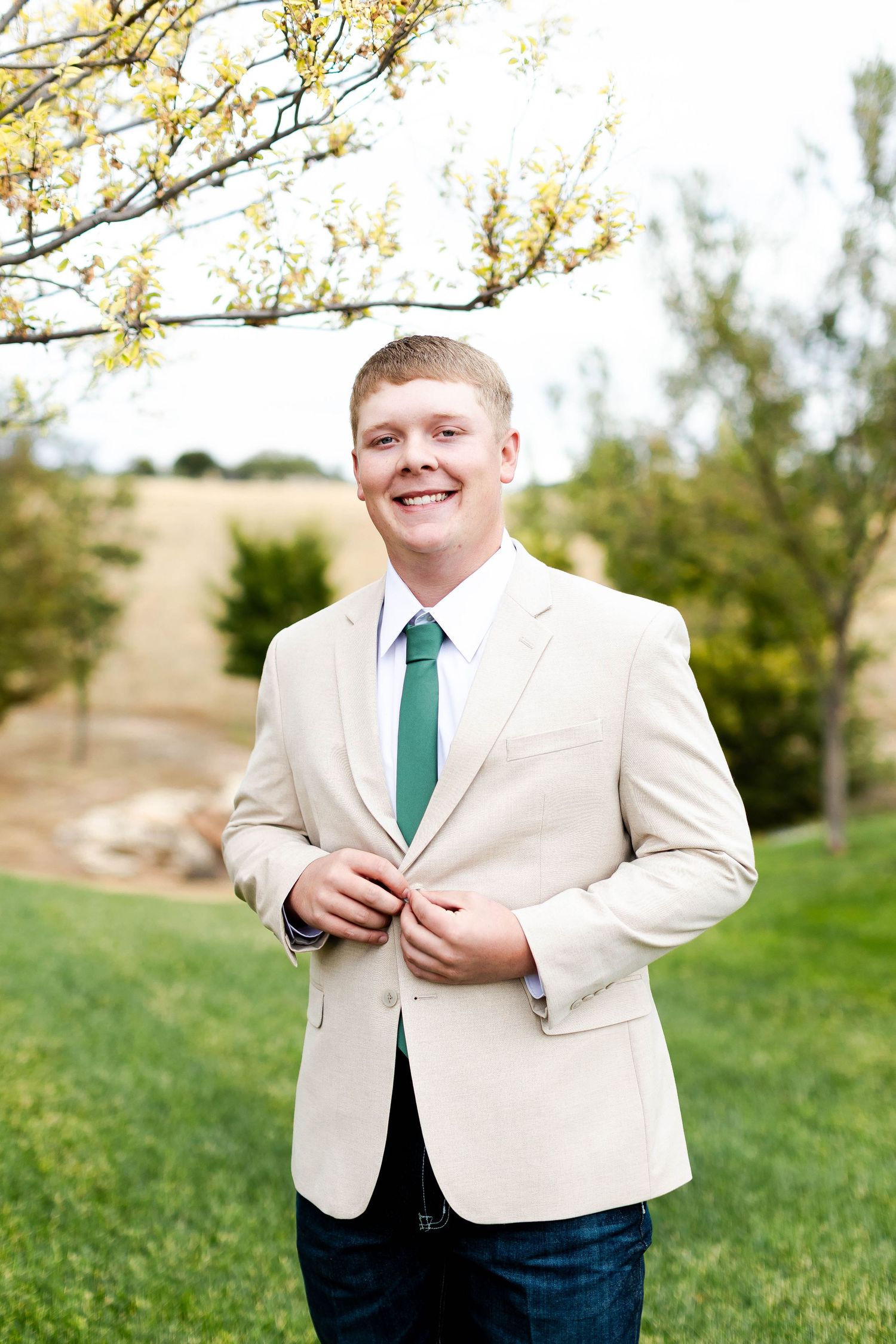 Person in a cream colored suit jacket and emerald green tie poses outdoors against a natural backdrop.