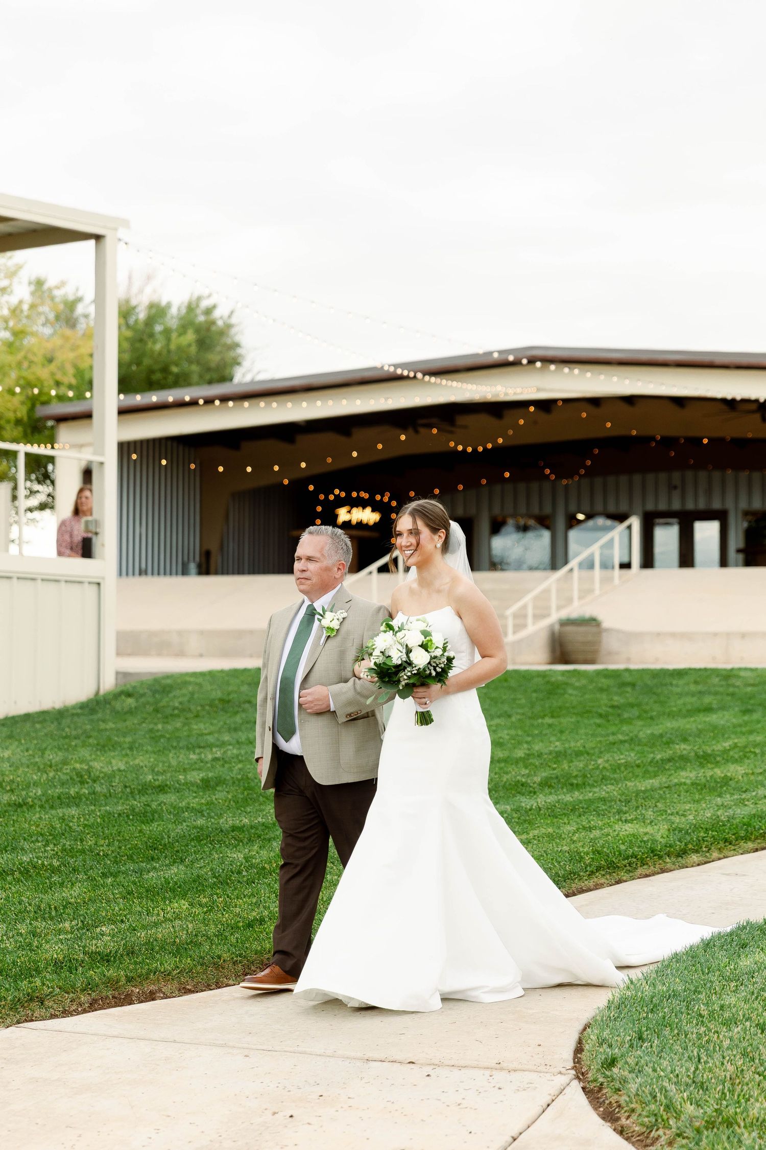 A wedding couple walks along a path outside a venue decorated with string lights at sunset.