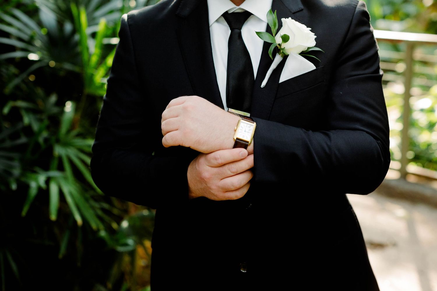 Close-up details of groom's formal attire with white boutonniere against greenery.