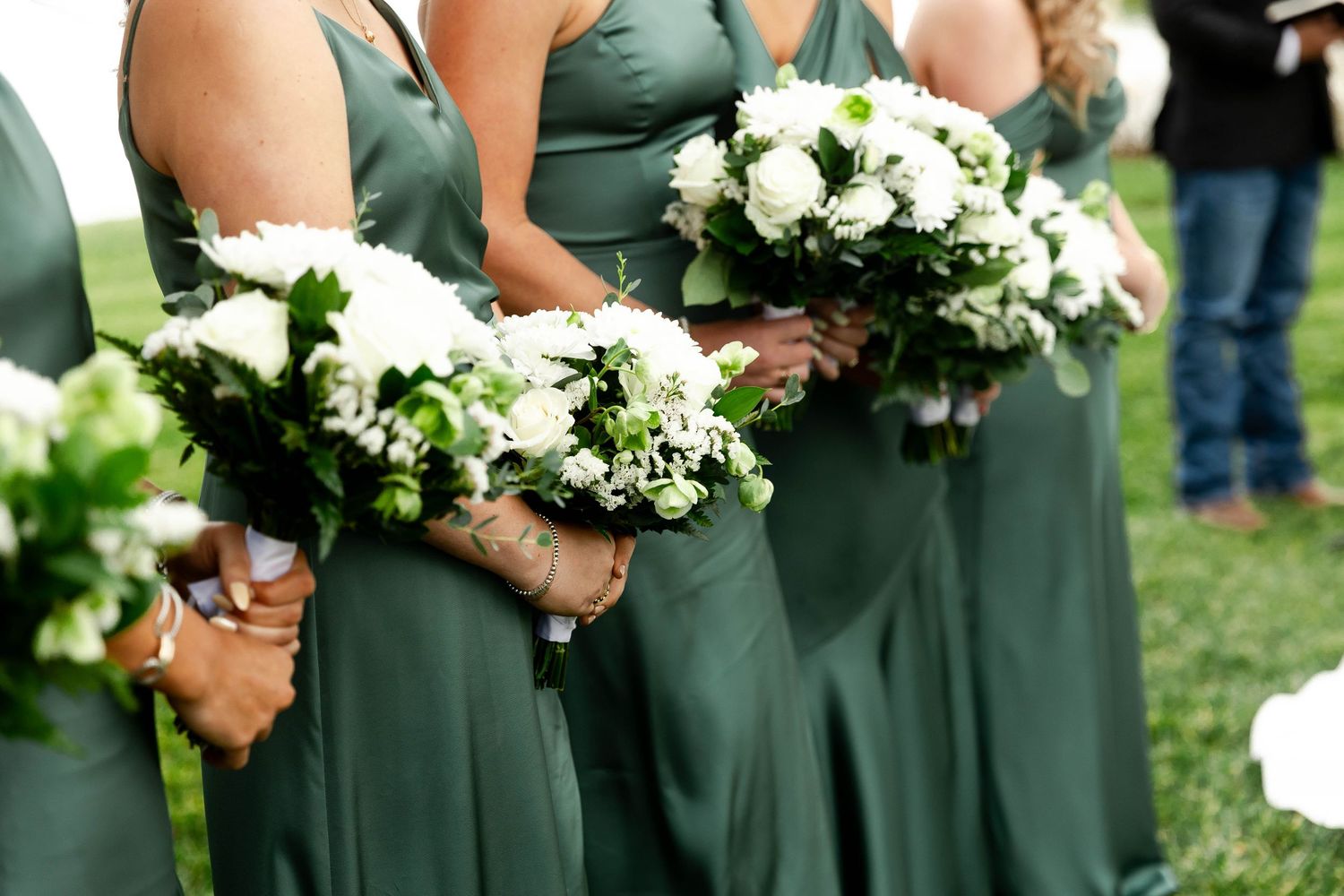 Bridesmaids in sage green dresses hold white floral bouquets during an outdoor ceremony.
