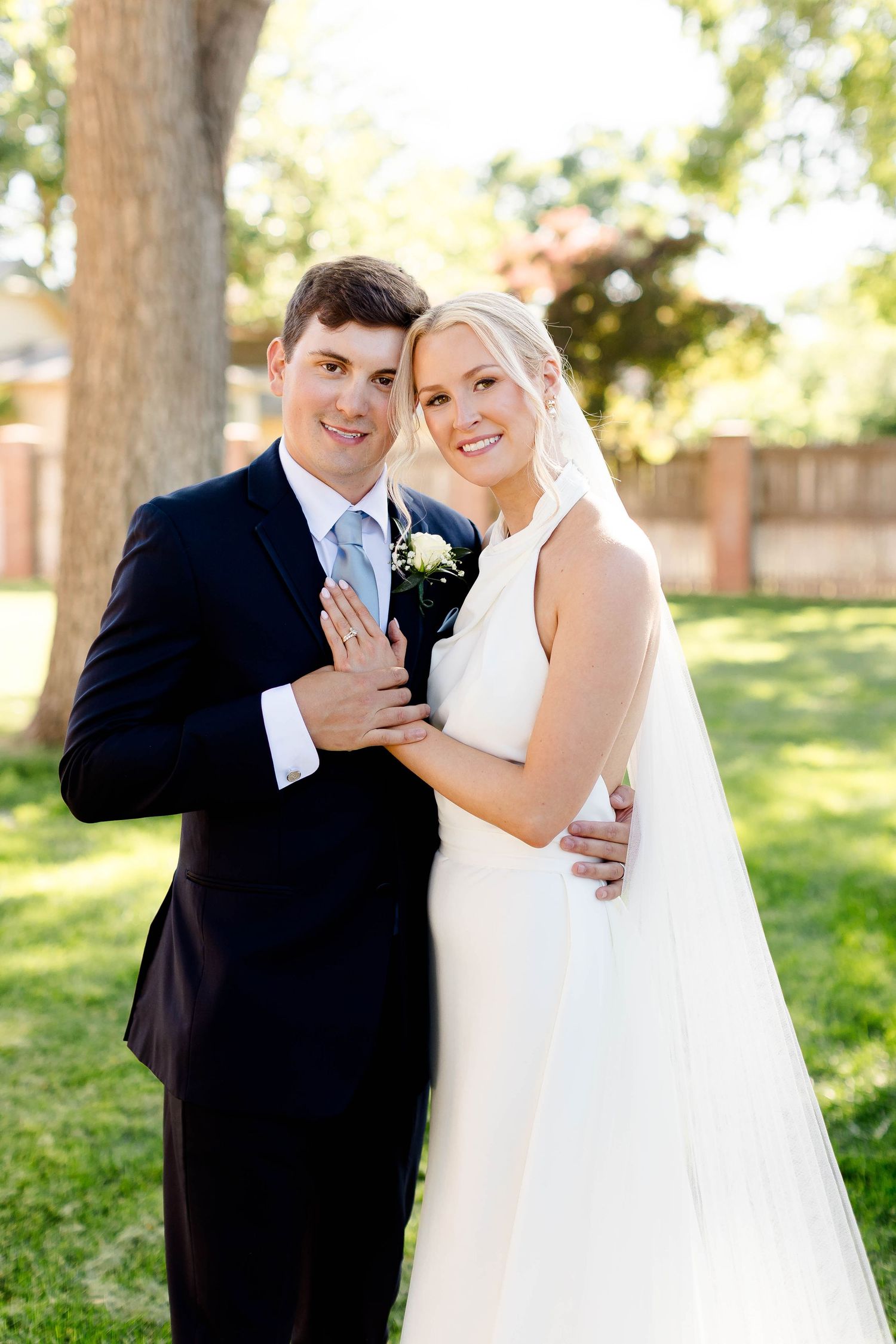 Joyful wedding portrait of couple in classic attire standing under a large tree.