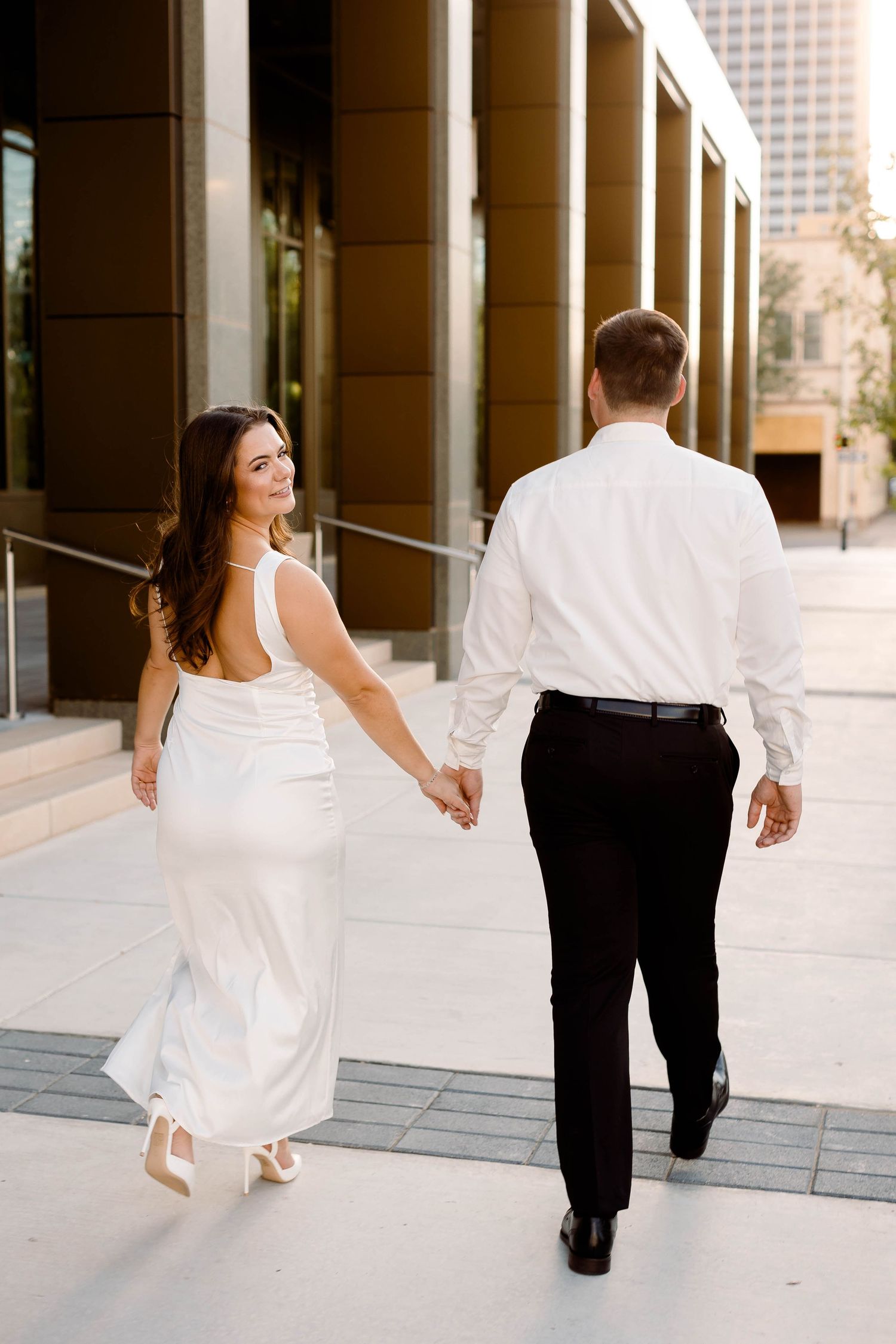 Couple in formal wear walking hand in hand along modern building exterior.