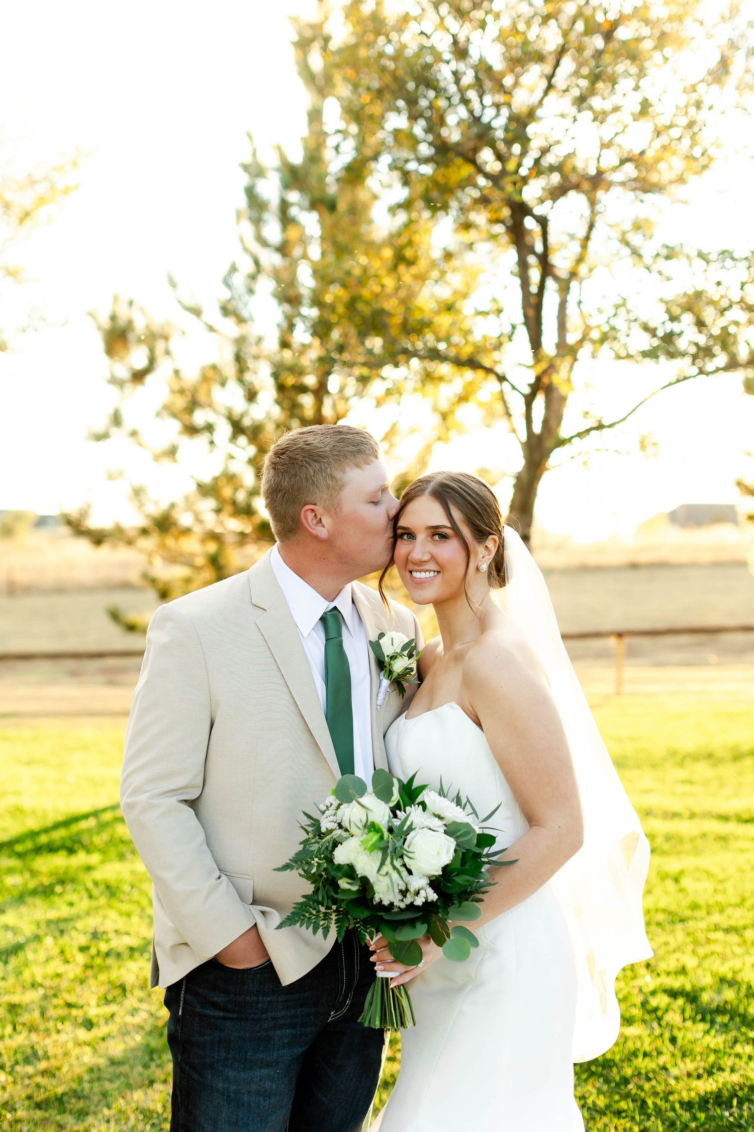 A couple in wedding attire embraces in a sunlit field at golden hour with white floral bouquet.