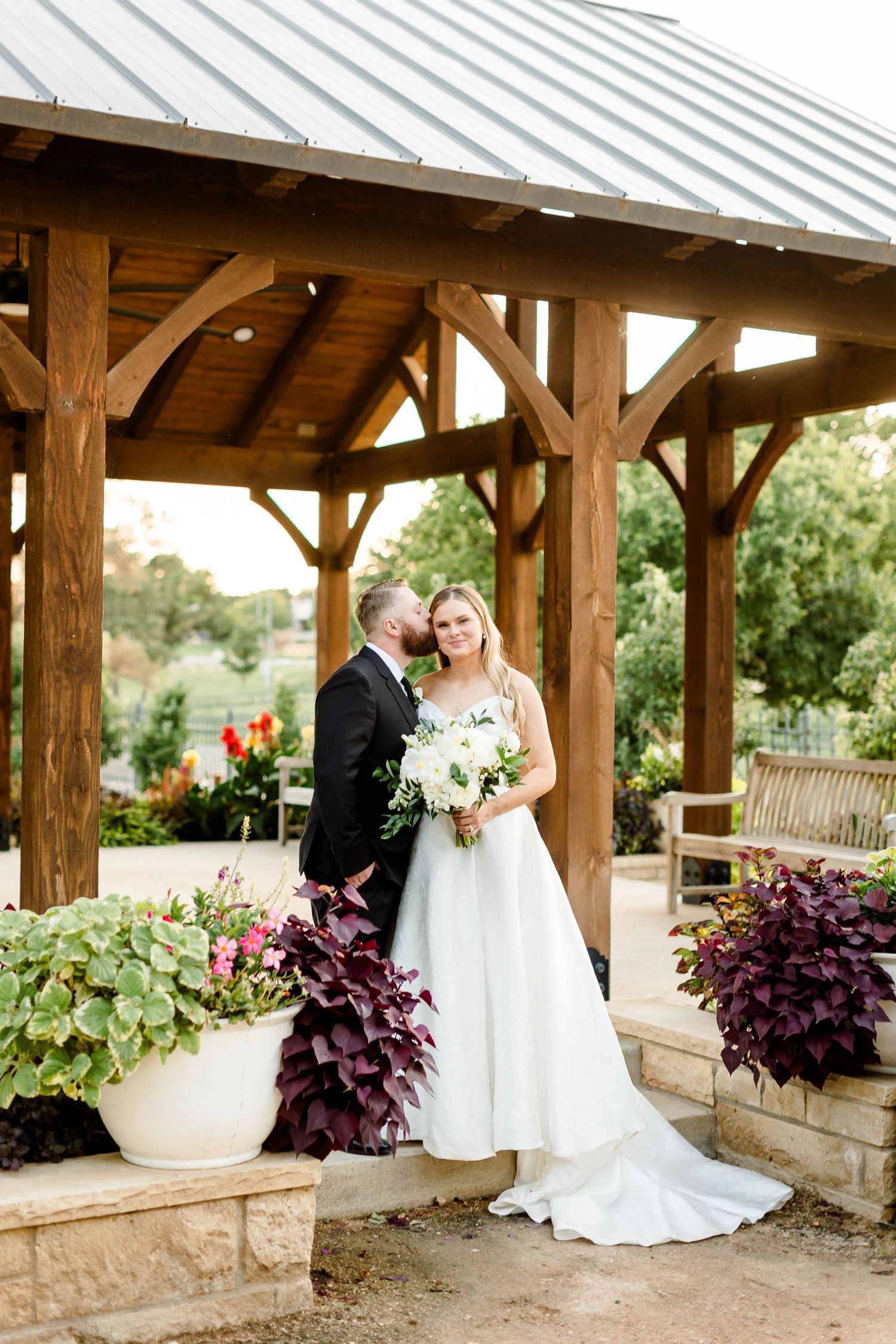Wedding portraits under a rustic wooden pavilion with stone columns and purple plants.
