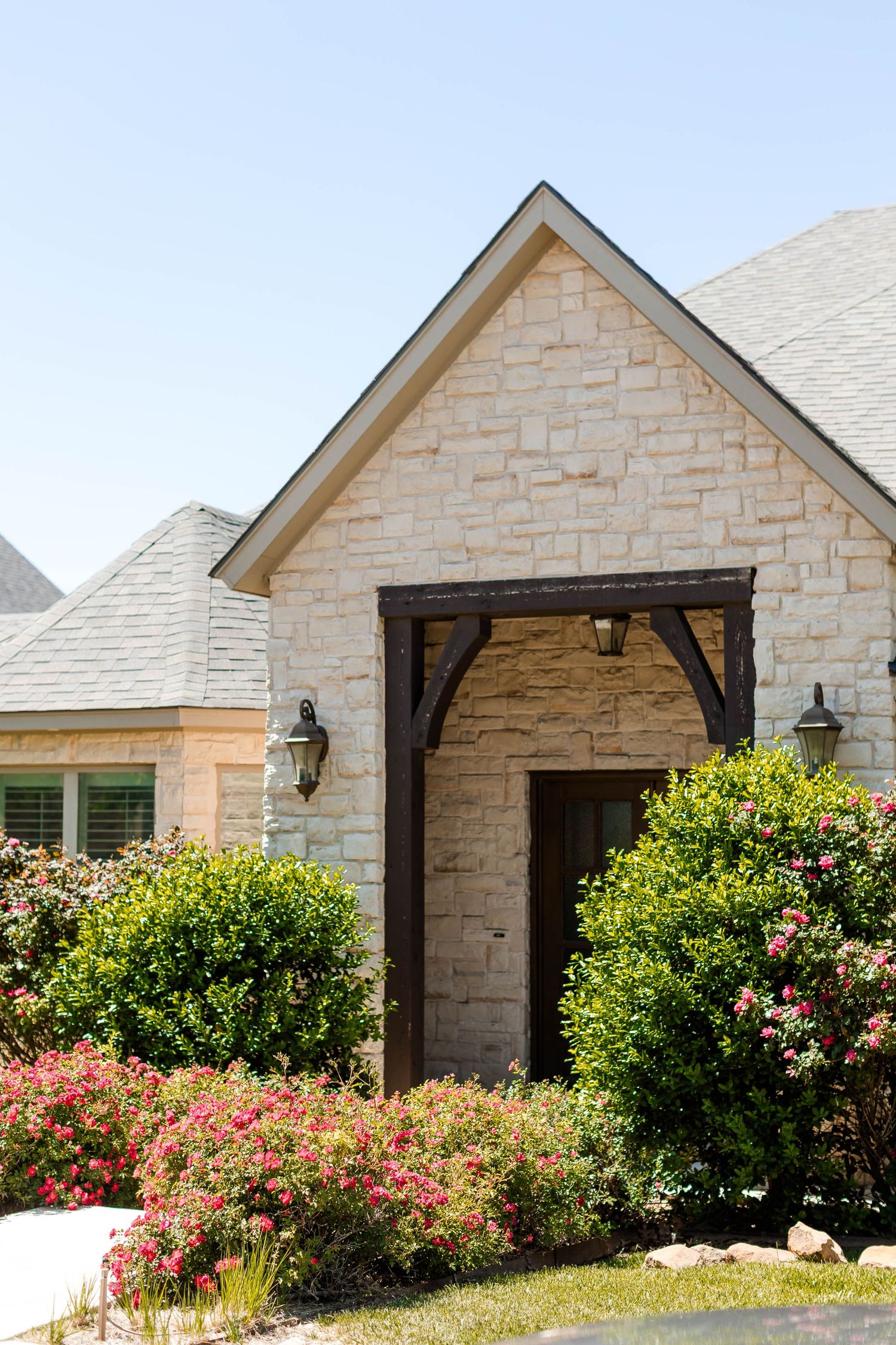 Stone facade of a modern house with blooming pink flowers and green shrubs in the landscaping.